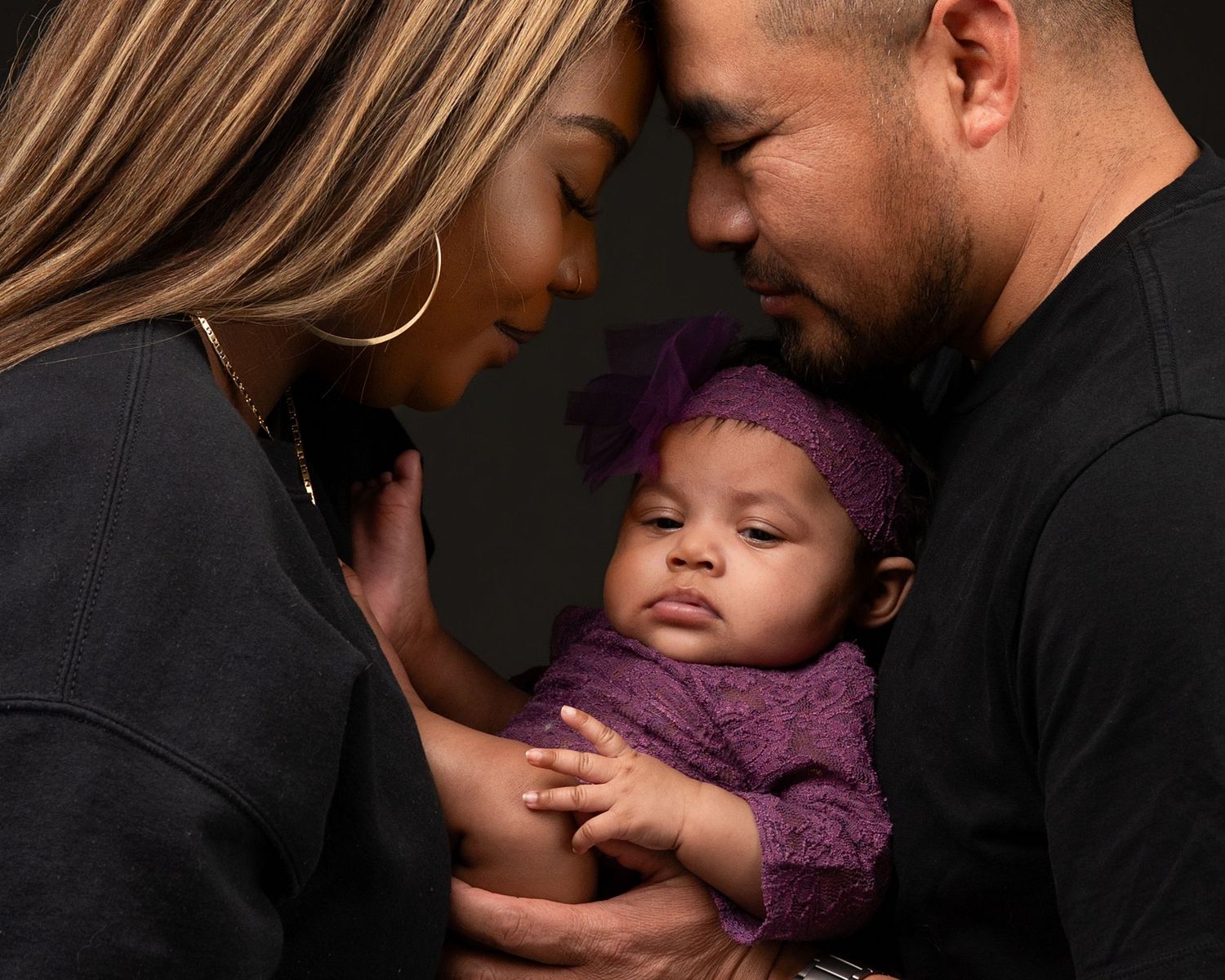 Loving parents tenderly hold their newborn baby wrapped in purple knit wrap in dark studio.