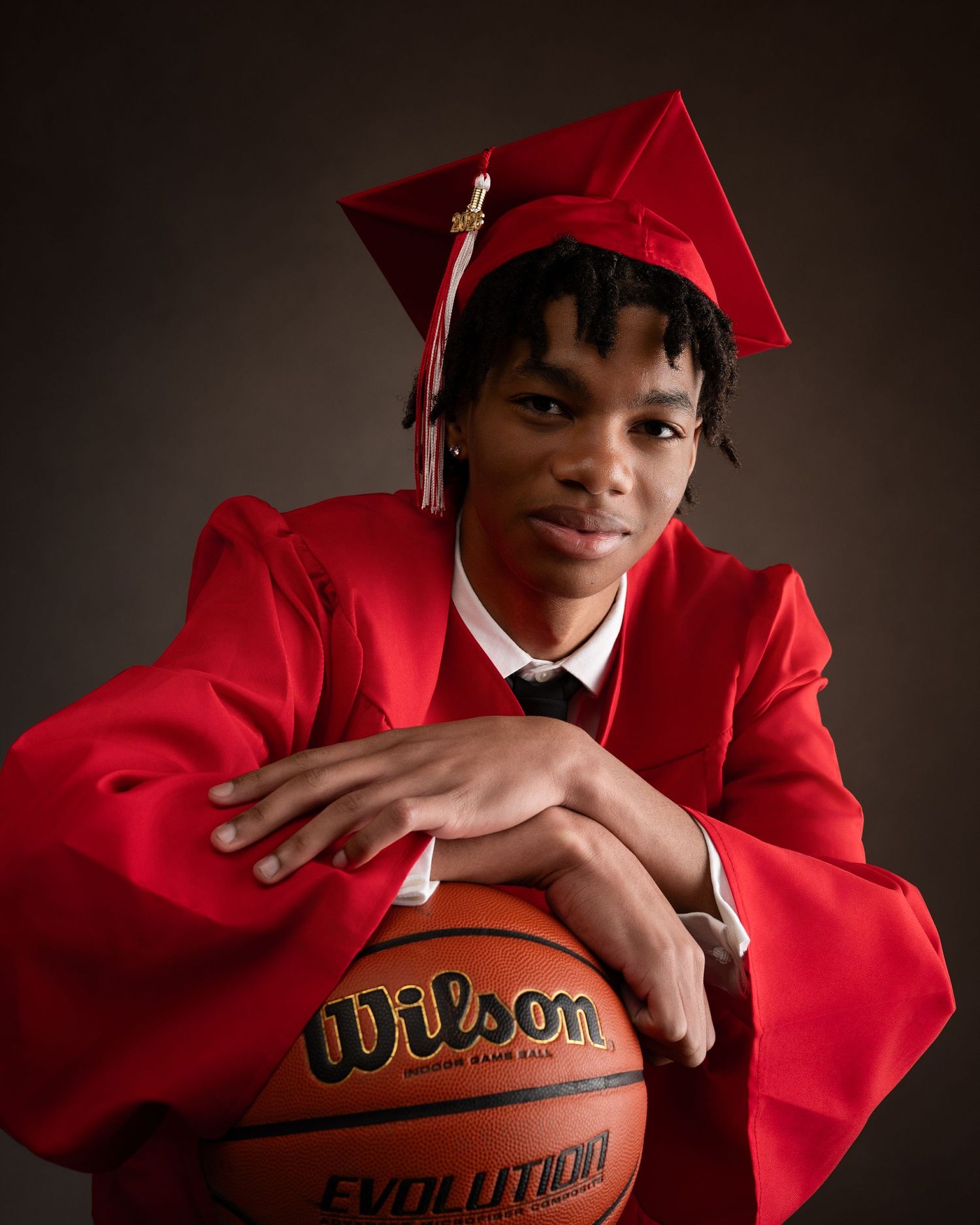 Senior student in red graduation cap and gown leans on Wilson Evolution basketball in dramatic portrait.