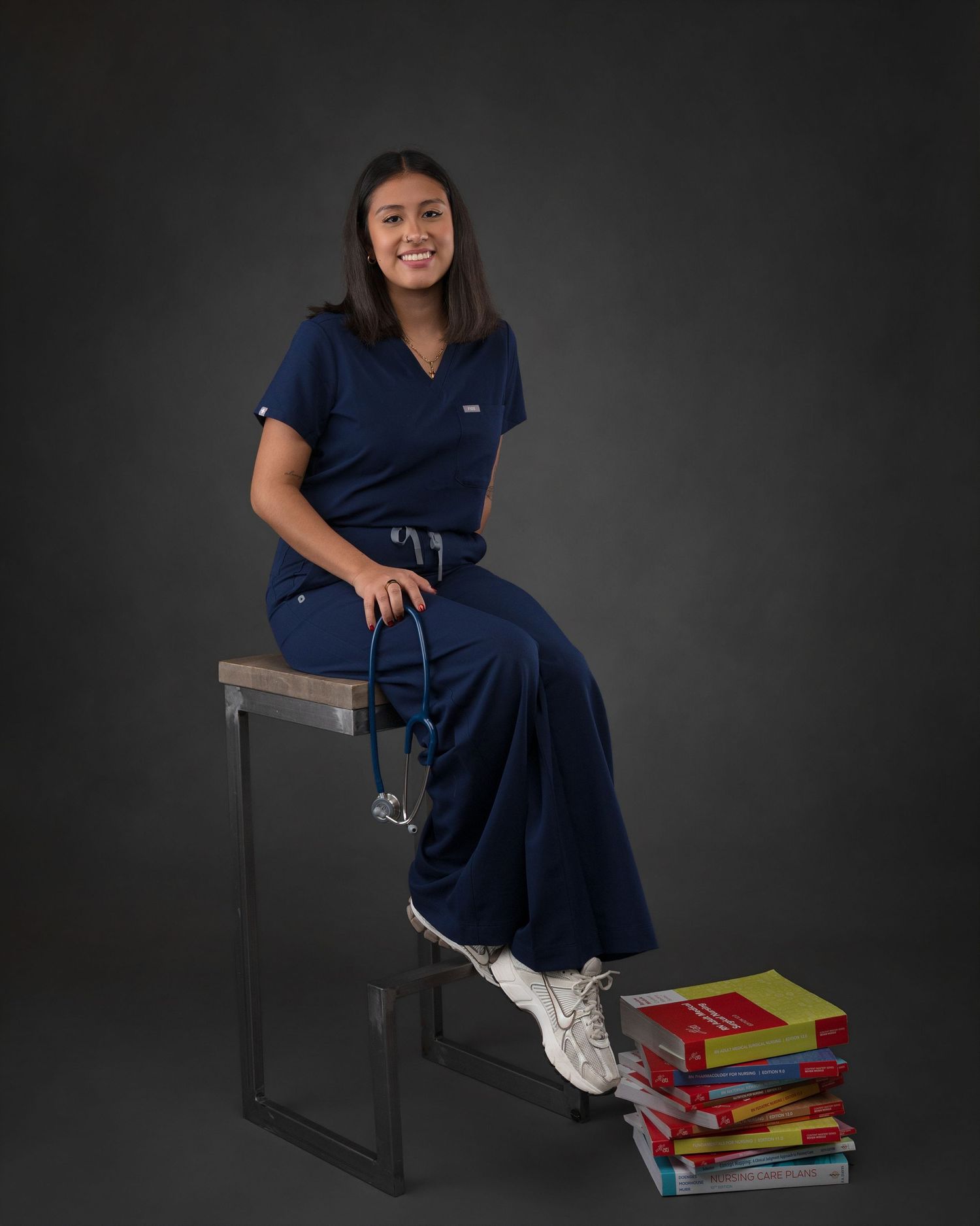 Young woman in navy blue scrubs sits on a stool with medical textbooks, nursing graduation photo.