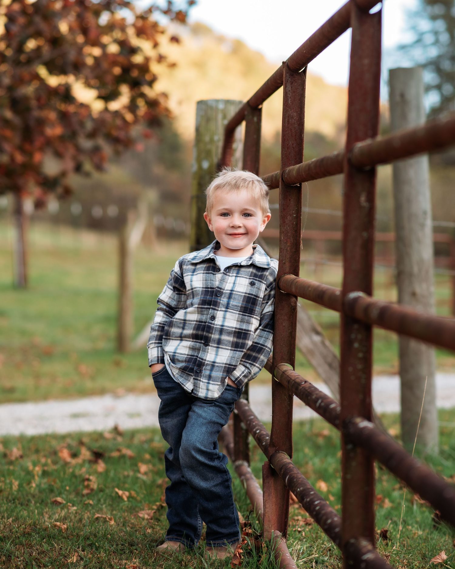 Happy young blonde boy in plaid flannel shirt leaning against rustic wooden fence in autumn outdoor setting.
