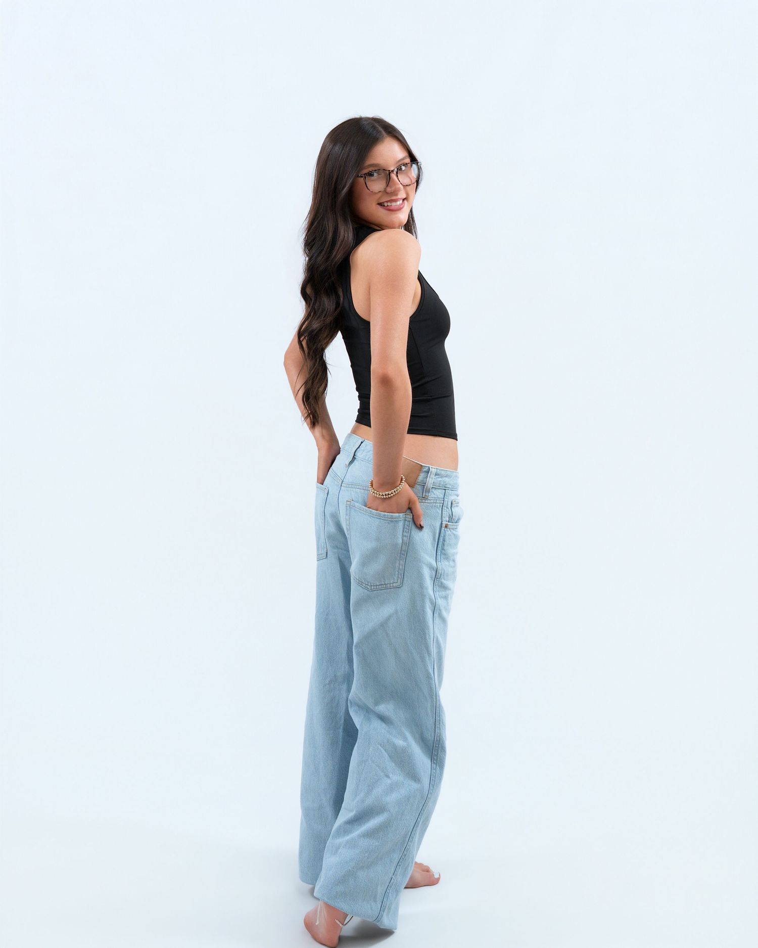 Young woman in black crop top and light blue wide-leg jeans posing sideways on white background.