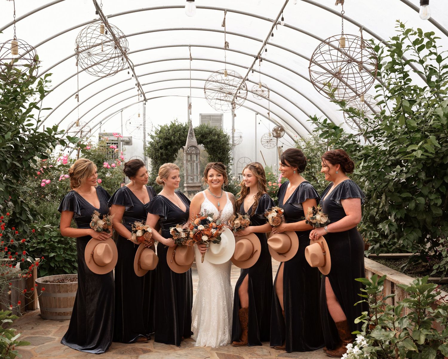 Bride with bridesmaids in navy dresses holding straw hats inside a lush greenhouse wedding venue setting.