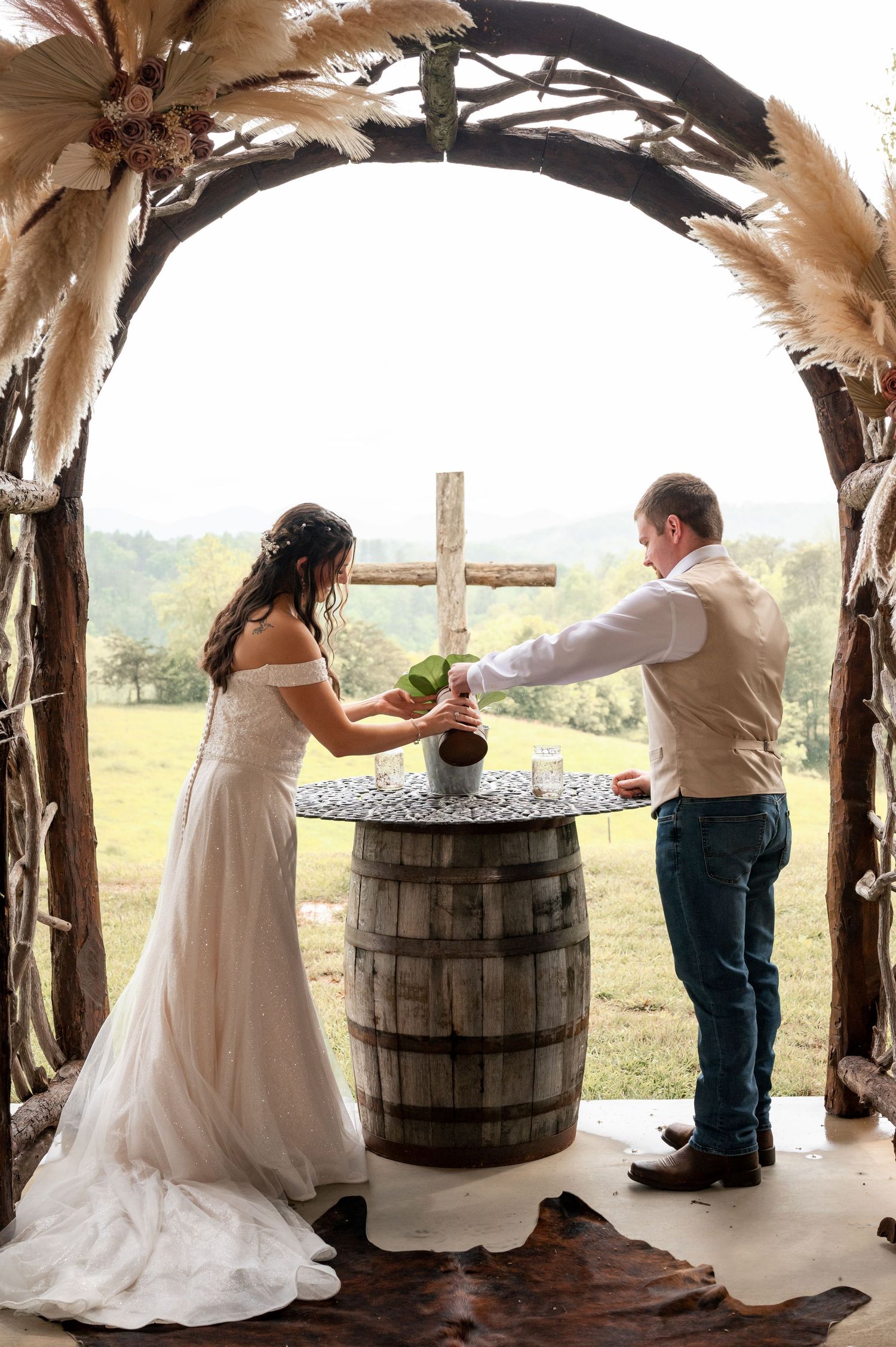Bride and groom performing a unity ceremony under a rustic wooden arch with a cross at an outdoor wedding.
