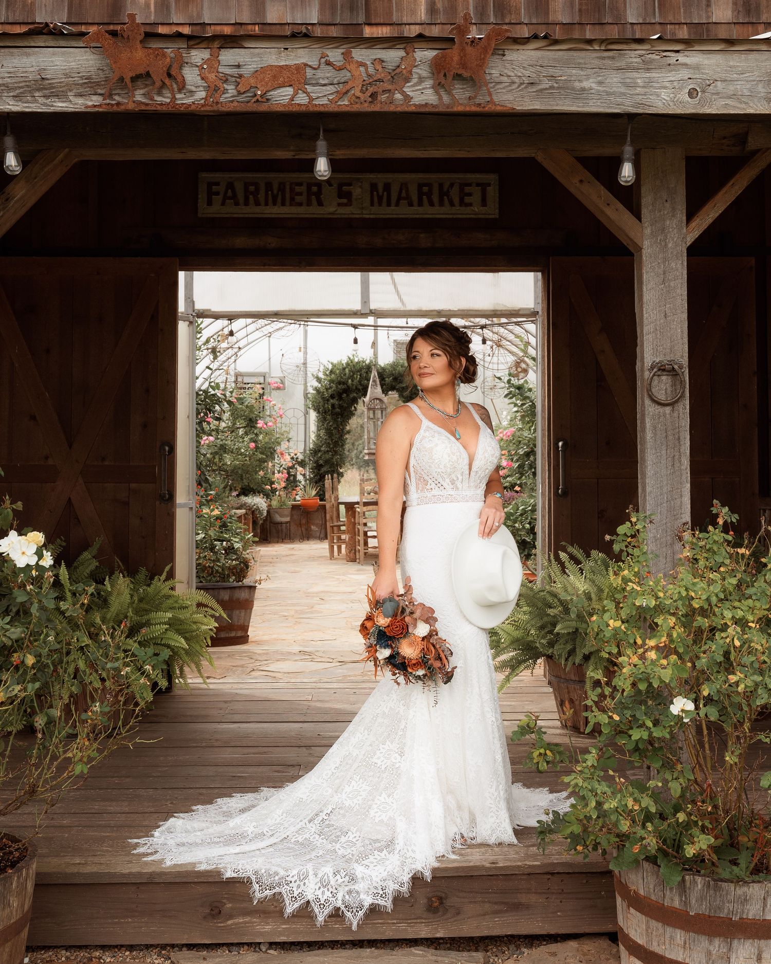 Bride in fitted lace gown with long train standing in rustic Farmer's Market venue with greenhouse backdrop.