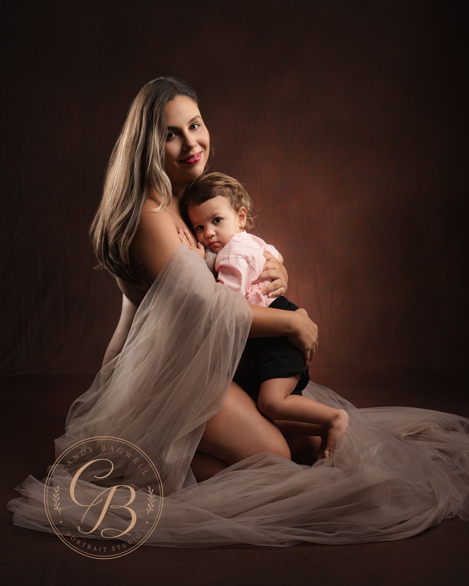 A mother in a sheer white gown sits gracefully holding her baby in a warm studio portrait.