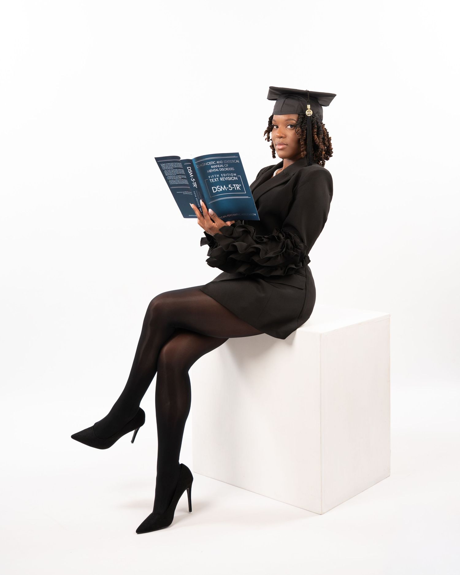 Graduate in black cap sitting on white cube reading DSM-5-TR book in studio.