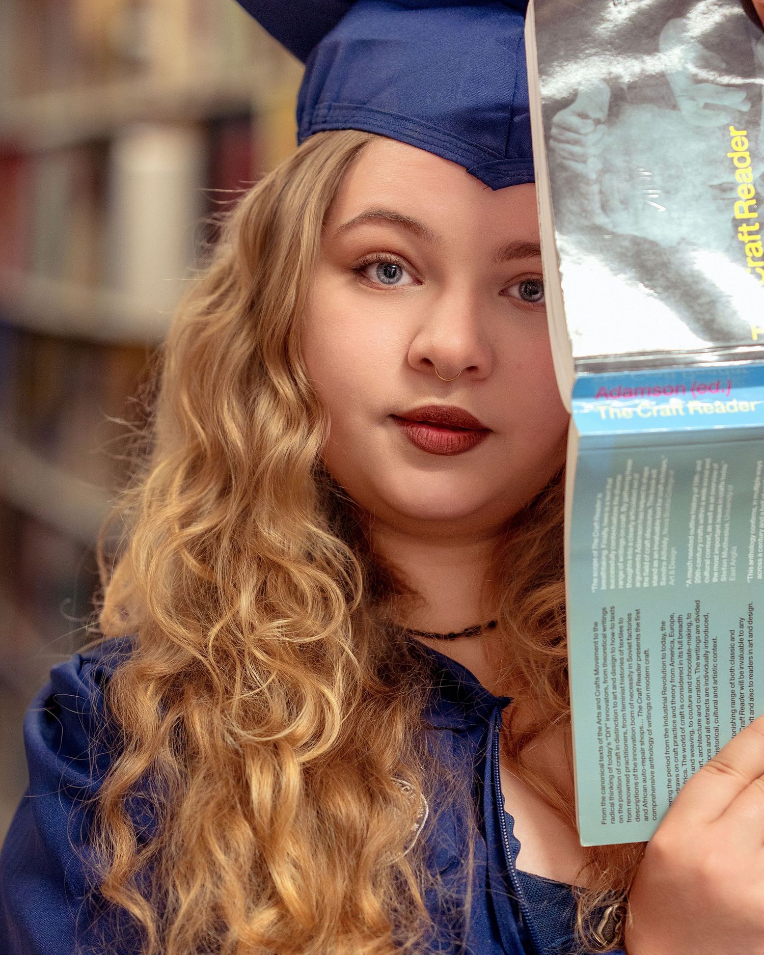 Female graduate in blue cap and gown holding open book near face in library setting.