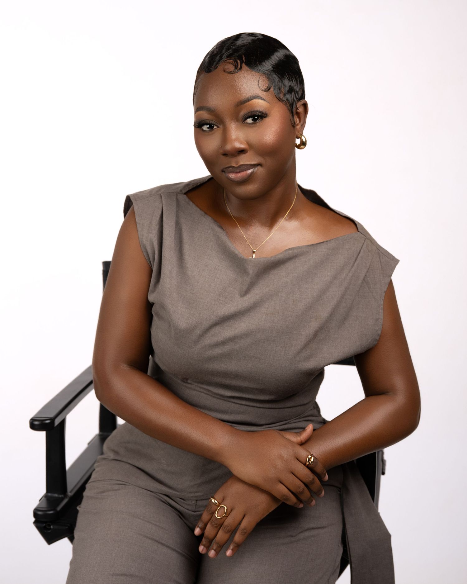 Professional woman in gray jumpsuit seated in director's chair, posing confidently against white background.