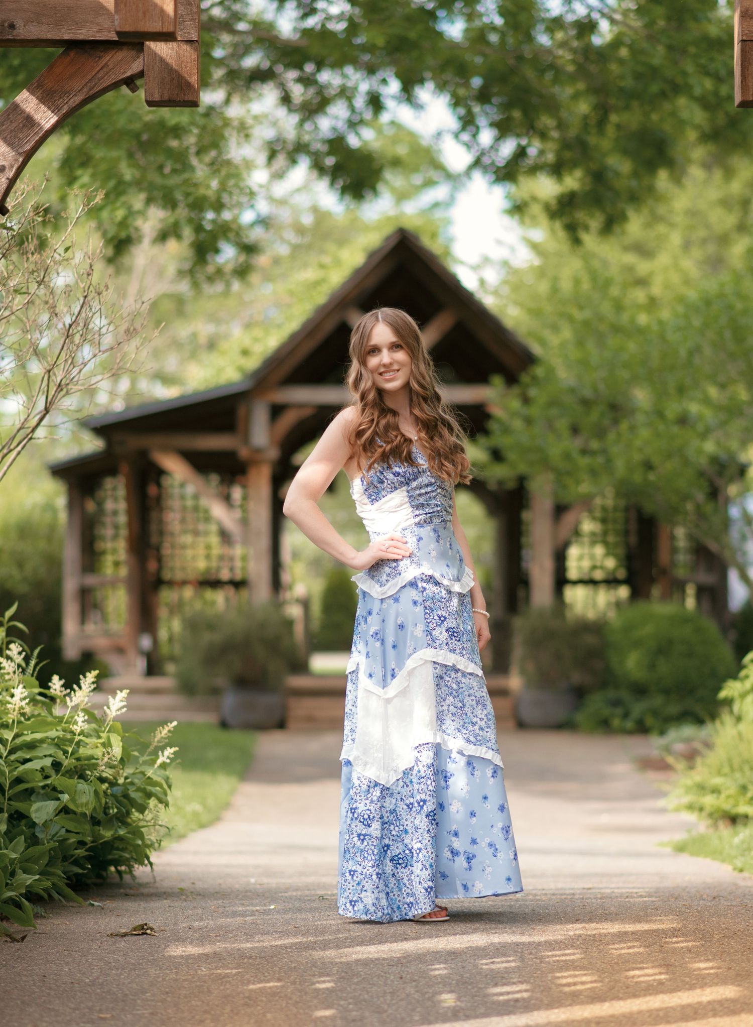 Beautiful young woman in blue floral maxi dress standing on garden path by gazebo.