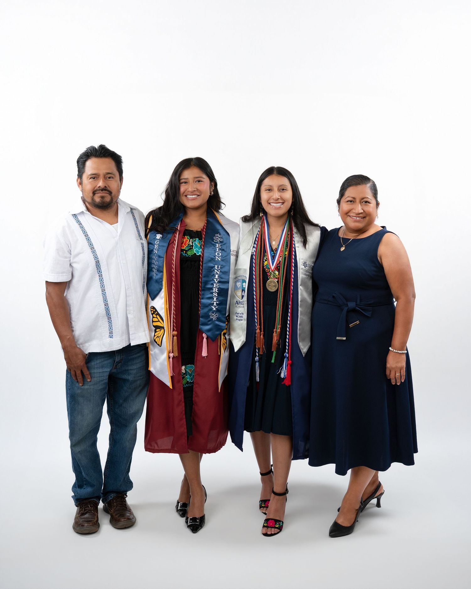 Family of four poses together with two graduates wearing colorful stoles in white studio portrait.