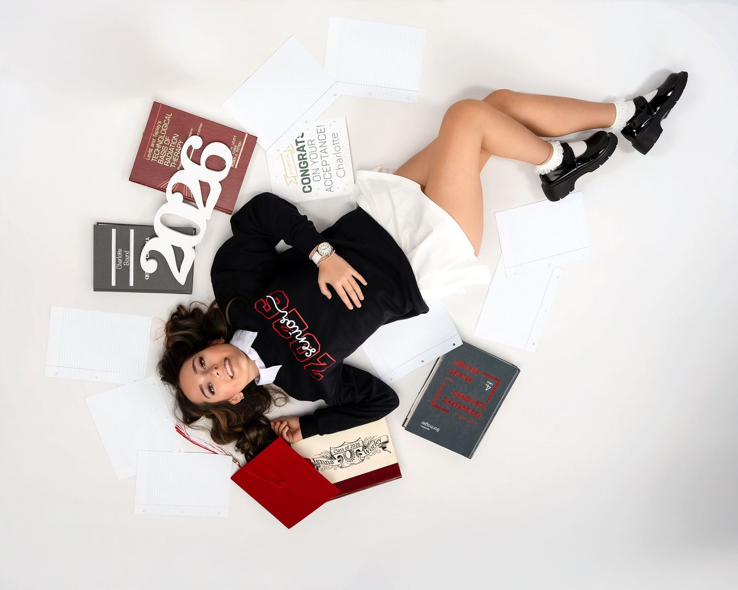 Graduate lies on white floor surrounded by books, papers, and 2026 decor in aerial graduation photo.