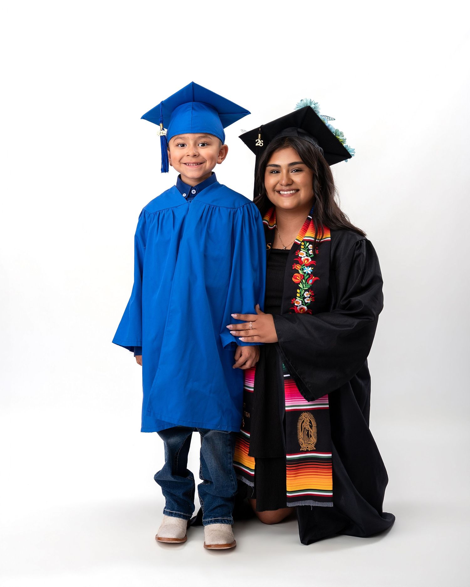 Young boy in blue cap and gown poses with female graduate in black gown and colorful cultural sash.