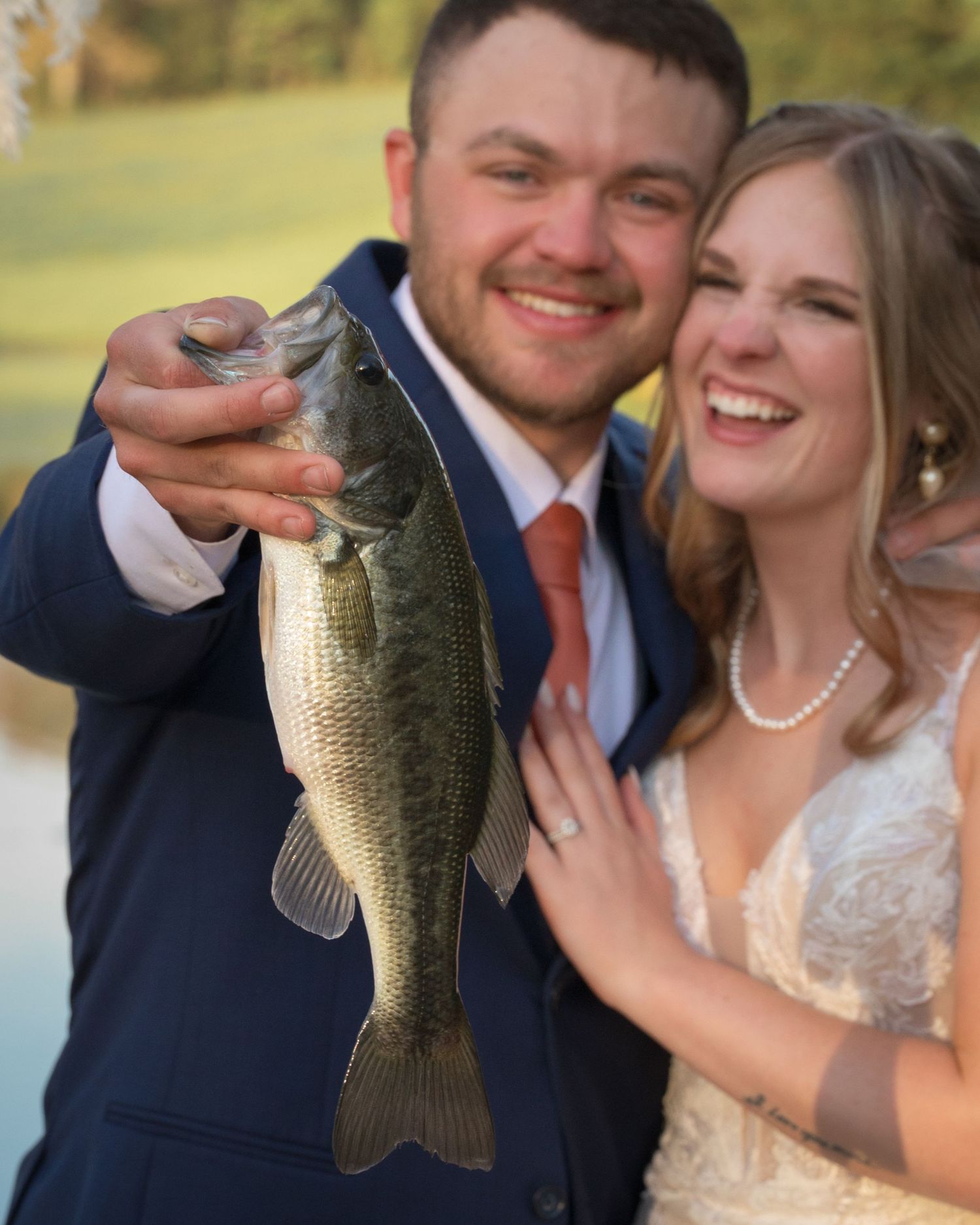 Smiling bride and groom in wedding attire proudly holding a large largemouth bass fish outdoors.