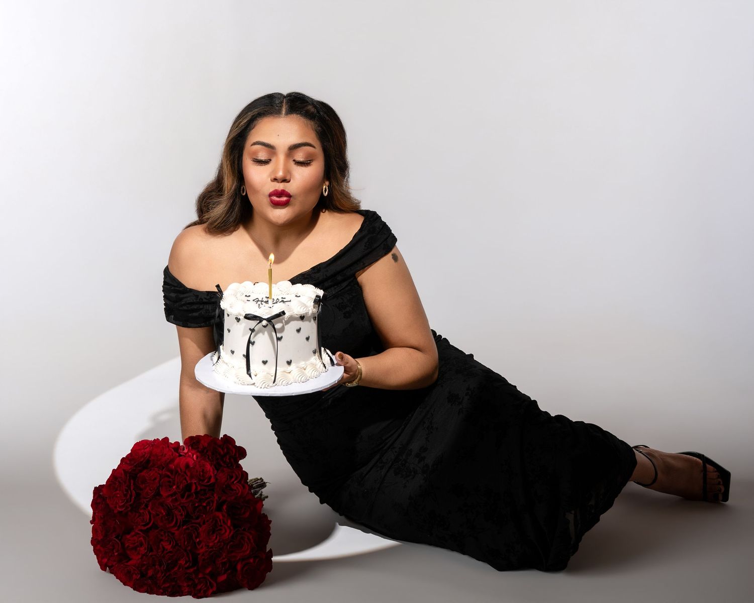 Woman in an off-shoulder black dress blowing out a candle on a small white cake beside a bouquet of red roses in a studio.