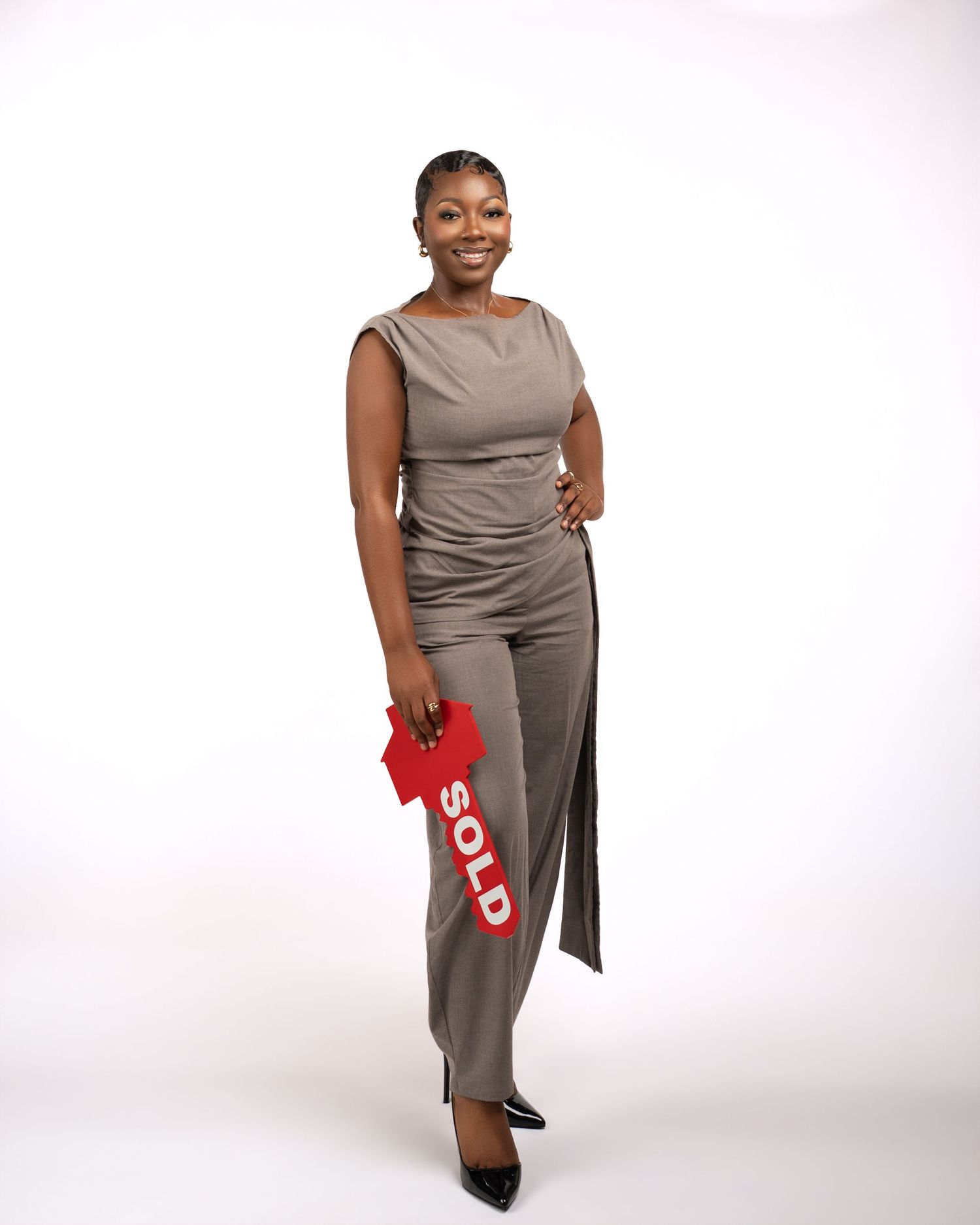 A professional woman in a gray jumpsuit holds a red SOLD real estate sign, posing confidently on a white background.