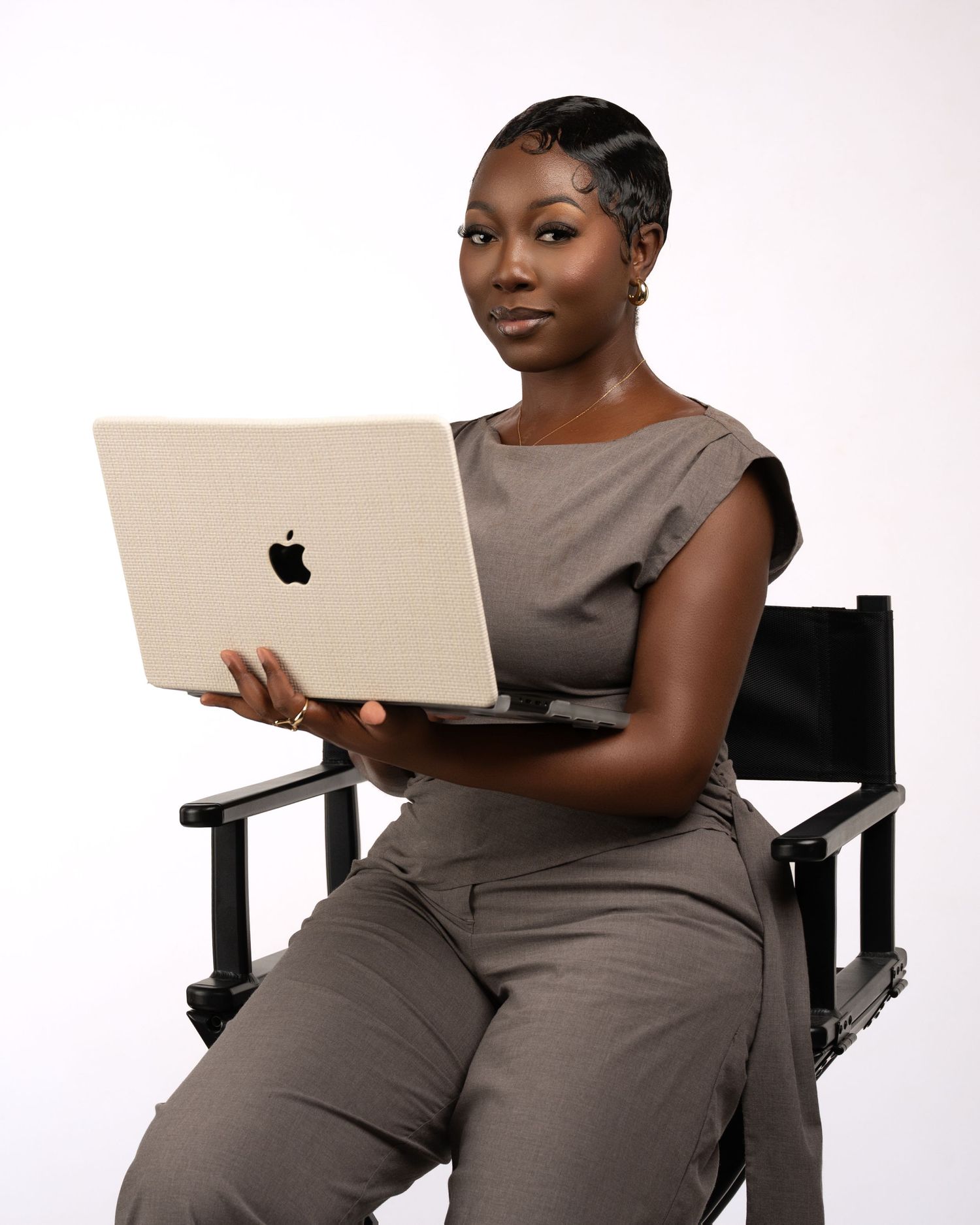 Professional woman in gray outfit holding Apple MacBook laptop while seated in director's chair on white background.