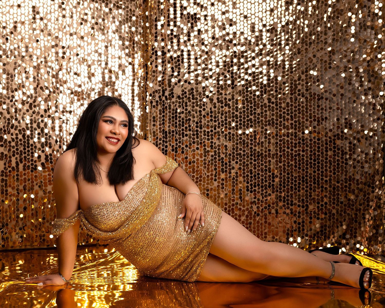 Woman in a sparkling gold sequin mini dress reclining on a reflective floor against a shimmering gold sequin backdrop.