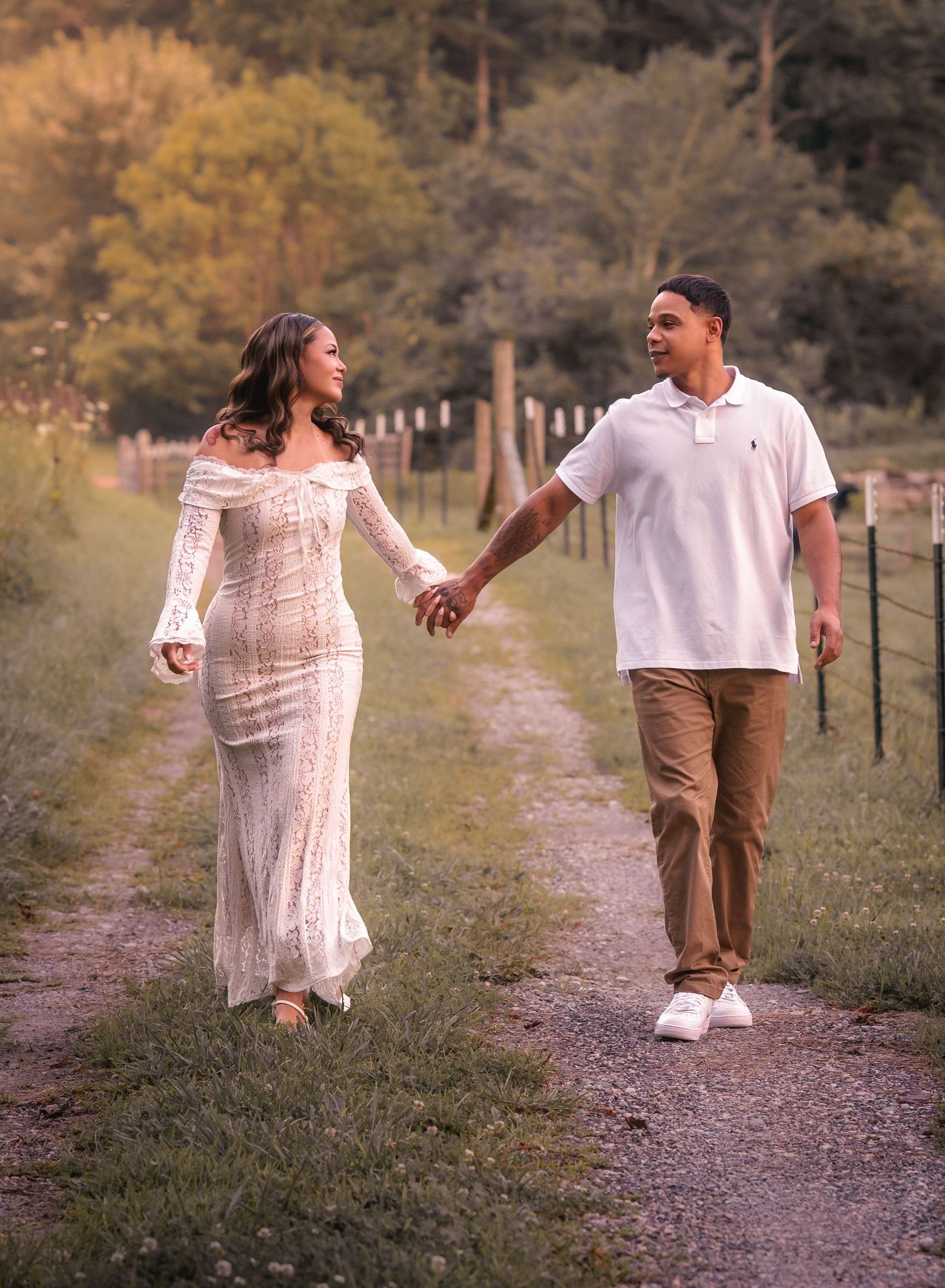 Couple walking hand-in-hand on a rustic dirt path, woman in white lace gown, man in white polo.
