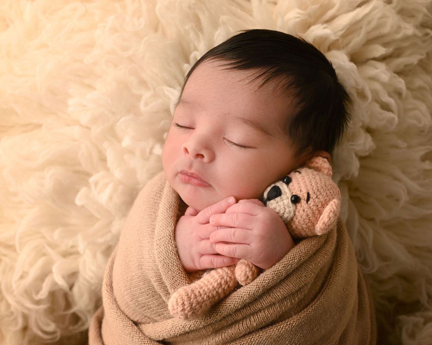 Sleeping newborn baby wrapped in a beige swaddle cuddling a small teddy bear on fluffy rug.