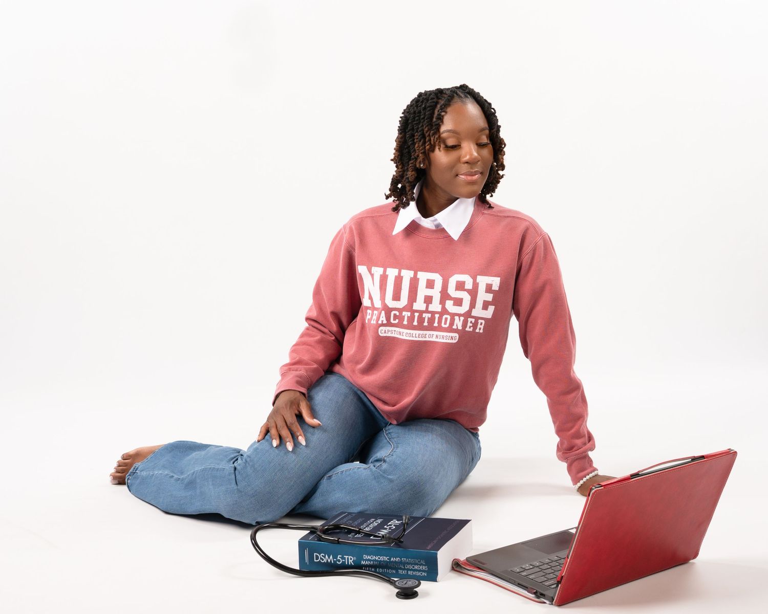 Woman in Nurse Practitioner sweatshirt sitting with laptop and blood pressure monitor.