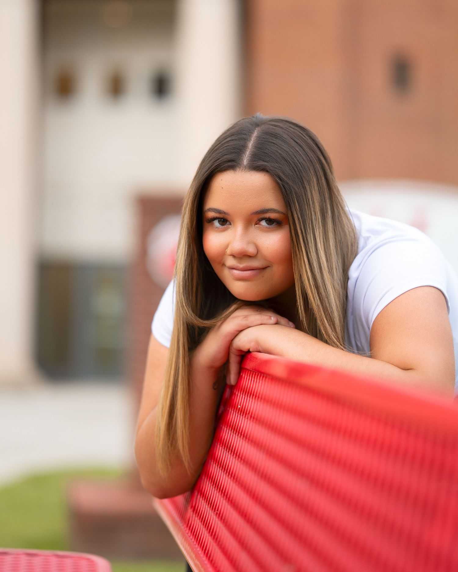 Young woman with highlighted hair leans on red chair smiling during outdoor senior portrait session.