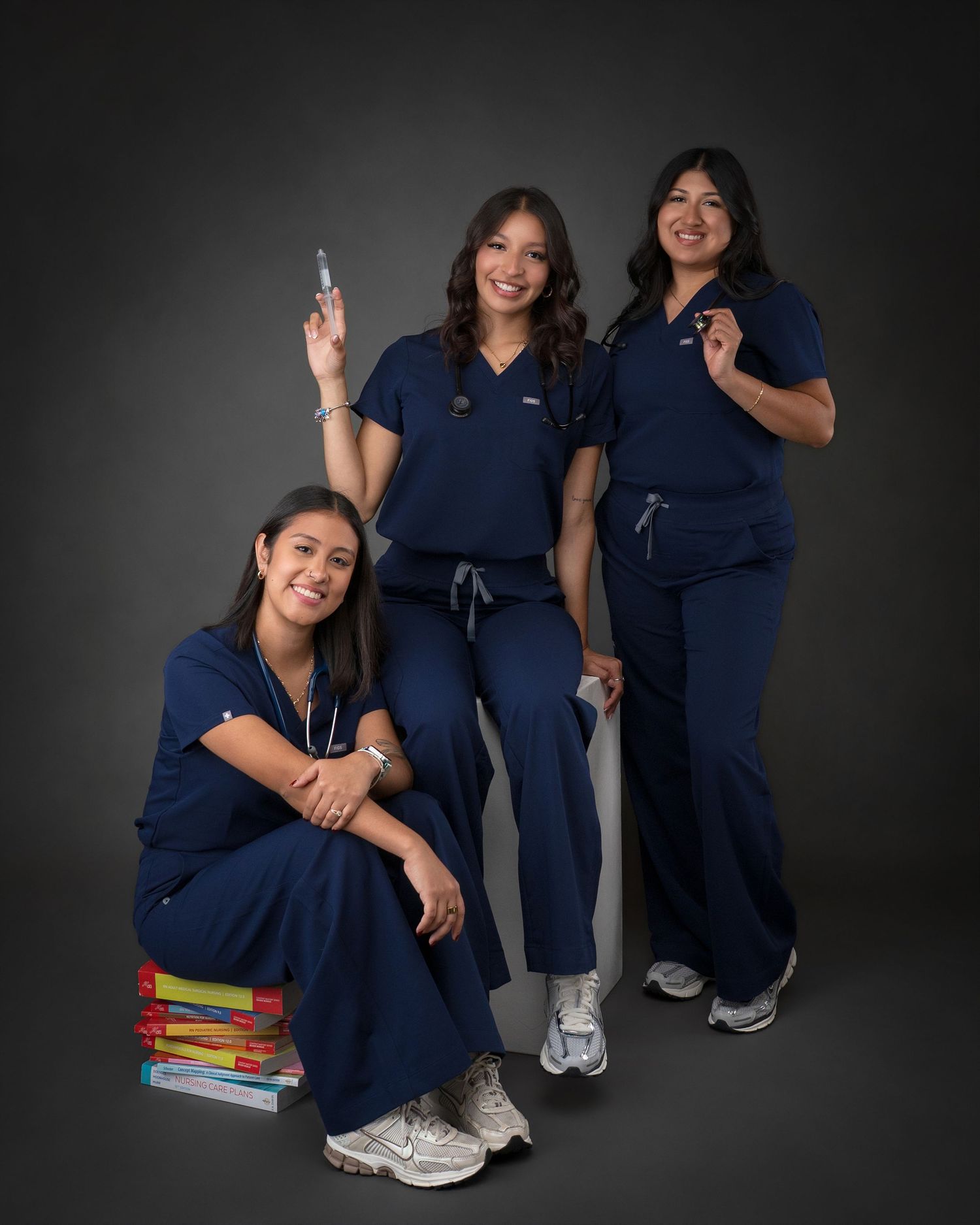 Three female nursing graduates pose together in navy scrubs with medical textbooks and a syringe.