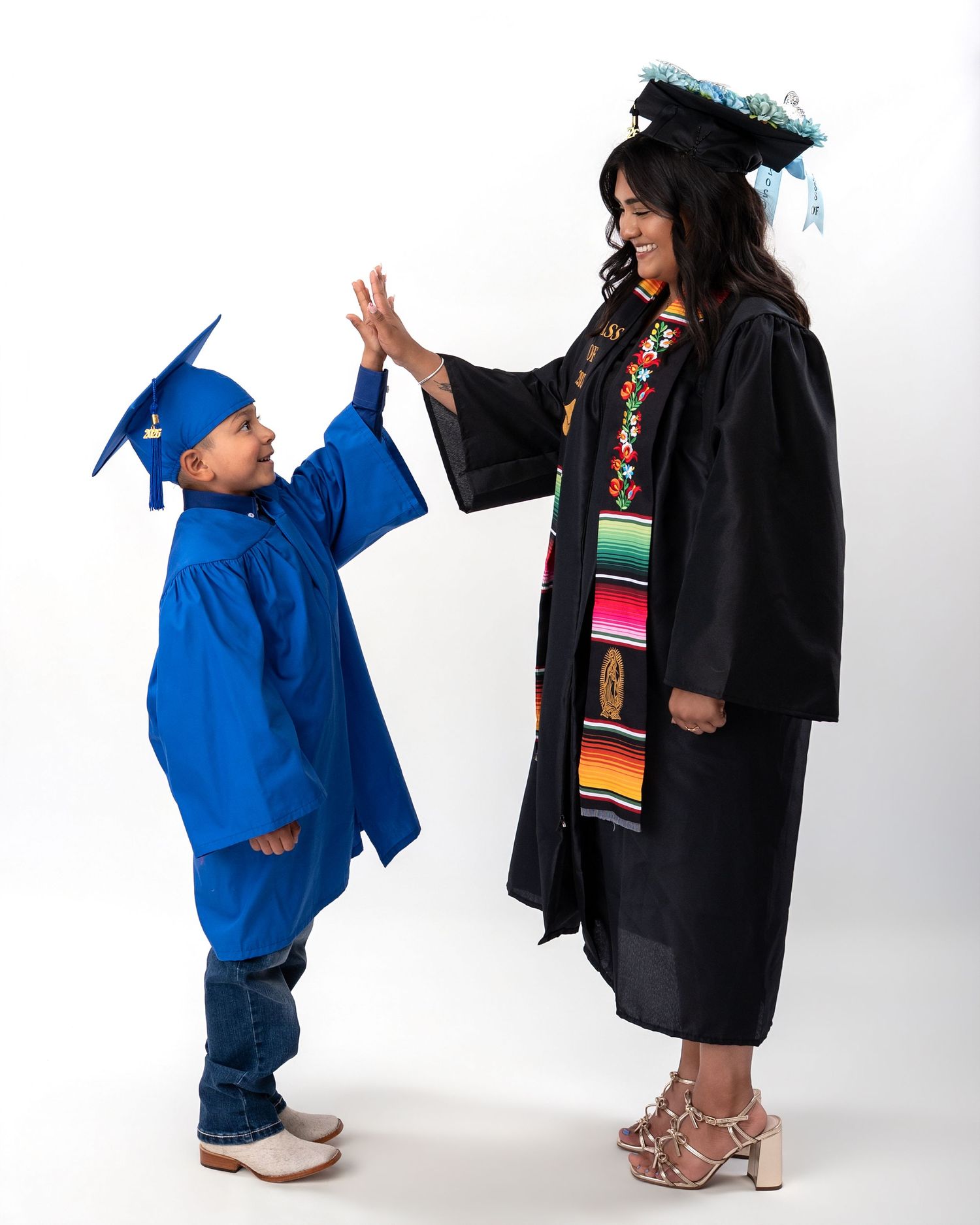 Young boy in blue cap and gown high-fives female graduate in black regalia in studio portrait.