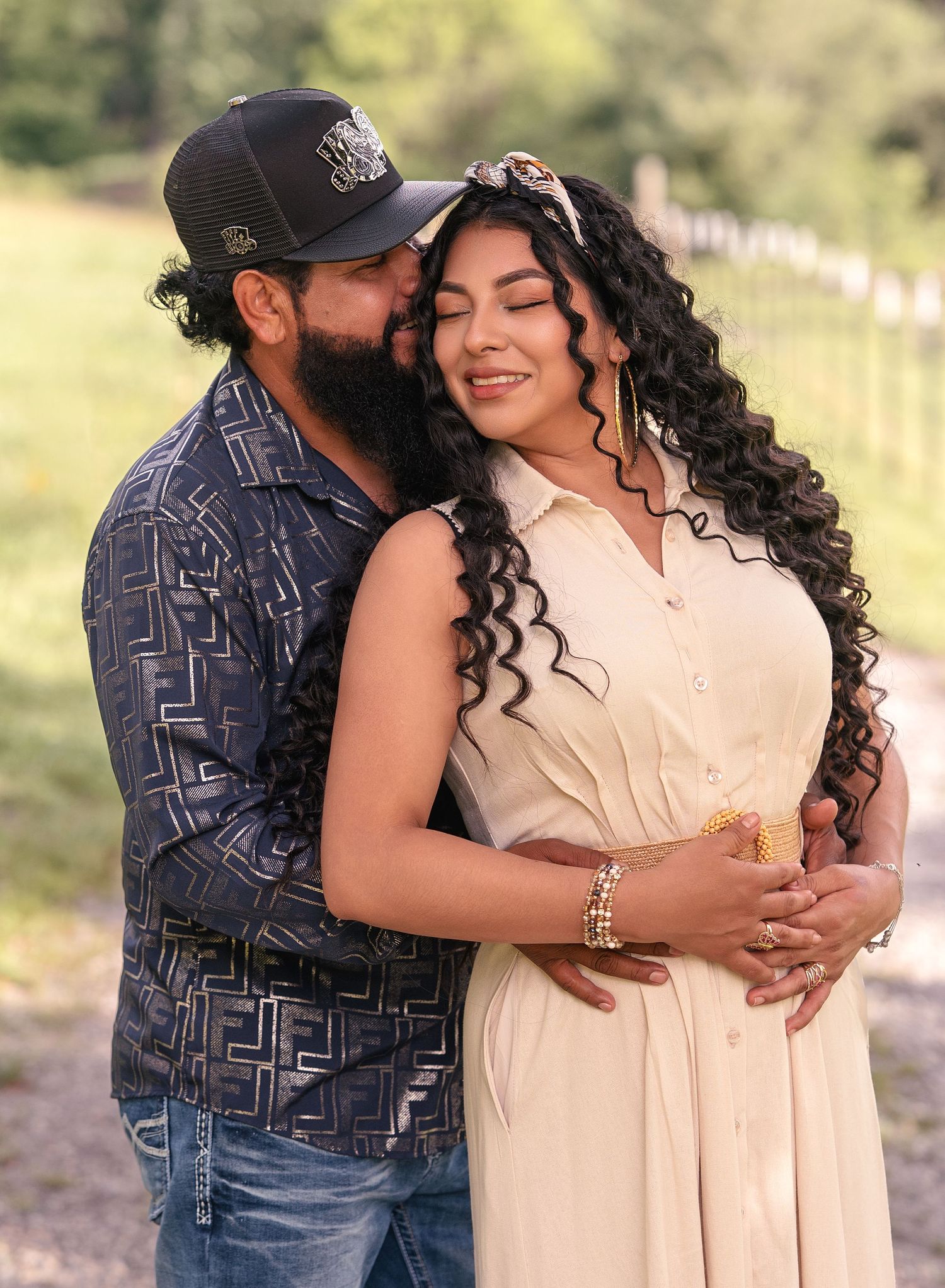 Couple embracing outdoors, man kissing womans forehead during maternity photo session in lush green setting.