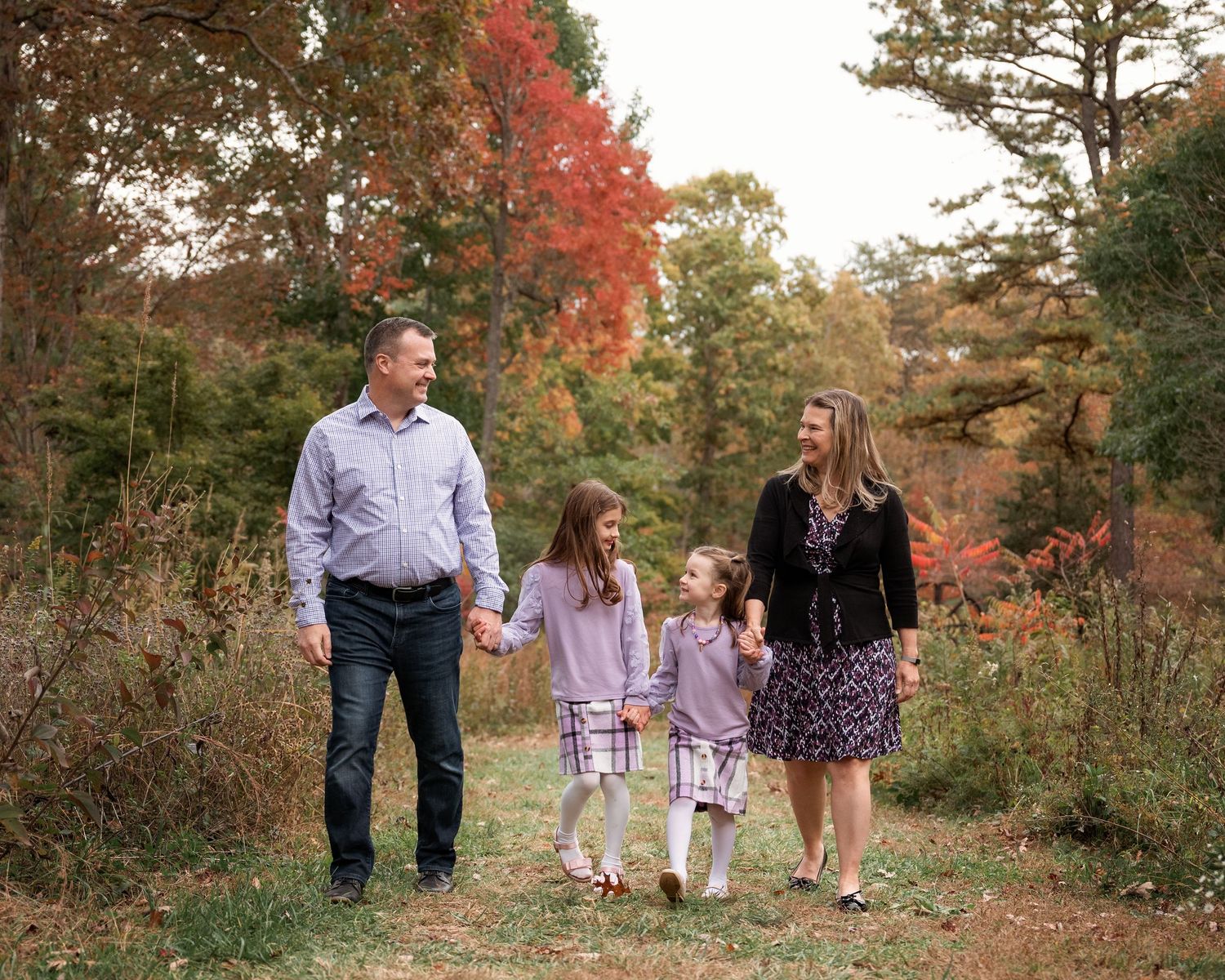 Happy family of four walking hand in hand through a scenic fall woodland path with vibrant red foliage.