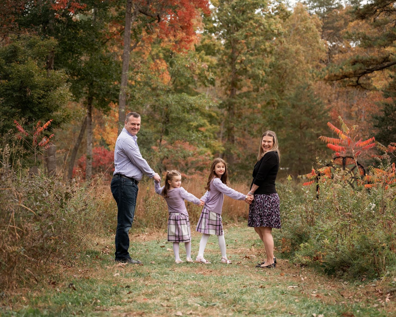 Family of four holding hands in a colorful autumn forest setting, parents and two girls in matching plaid dresses.