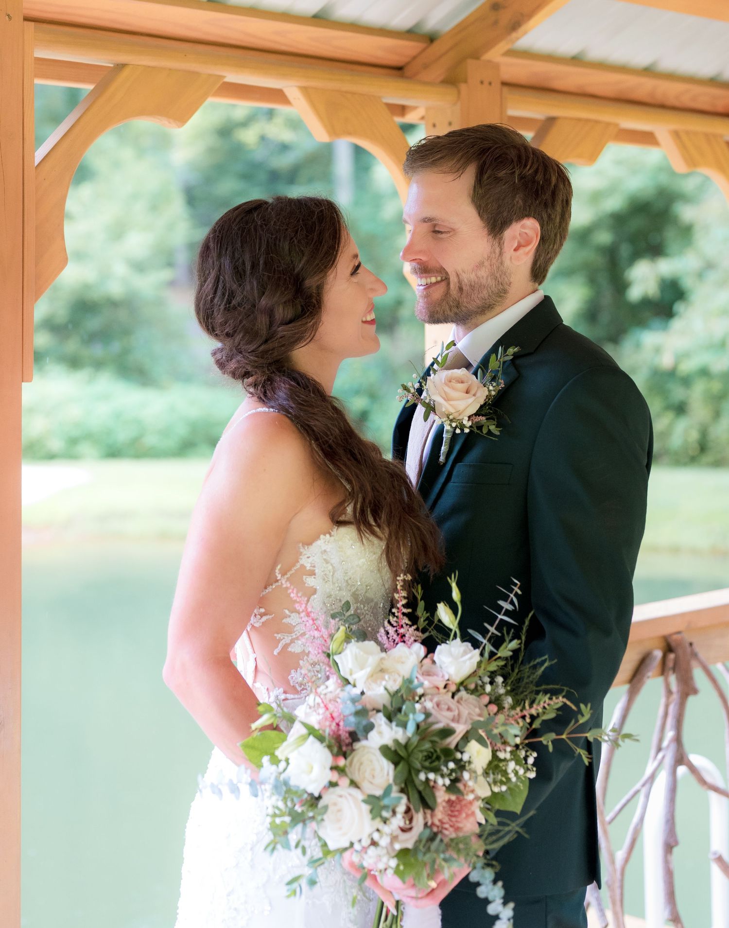 Smiling bride and groom share loving gaze under wooden pavilion at outdoor wedding.