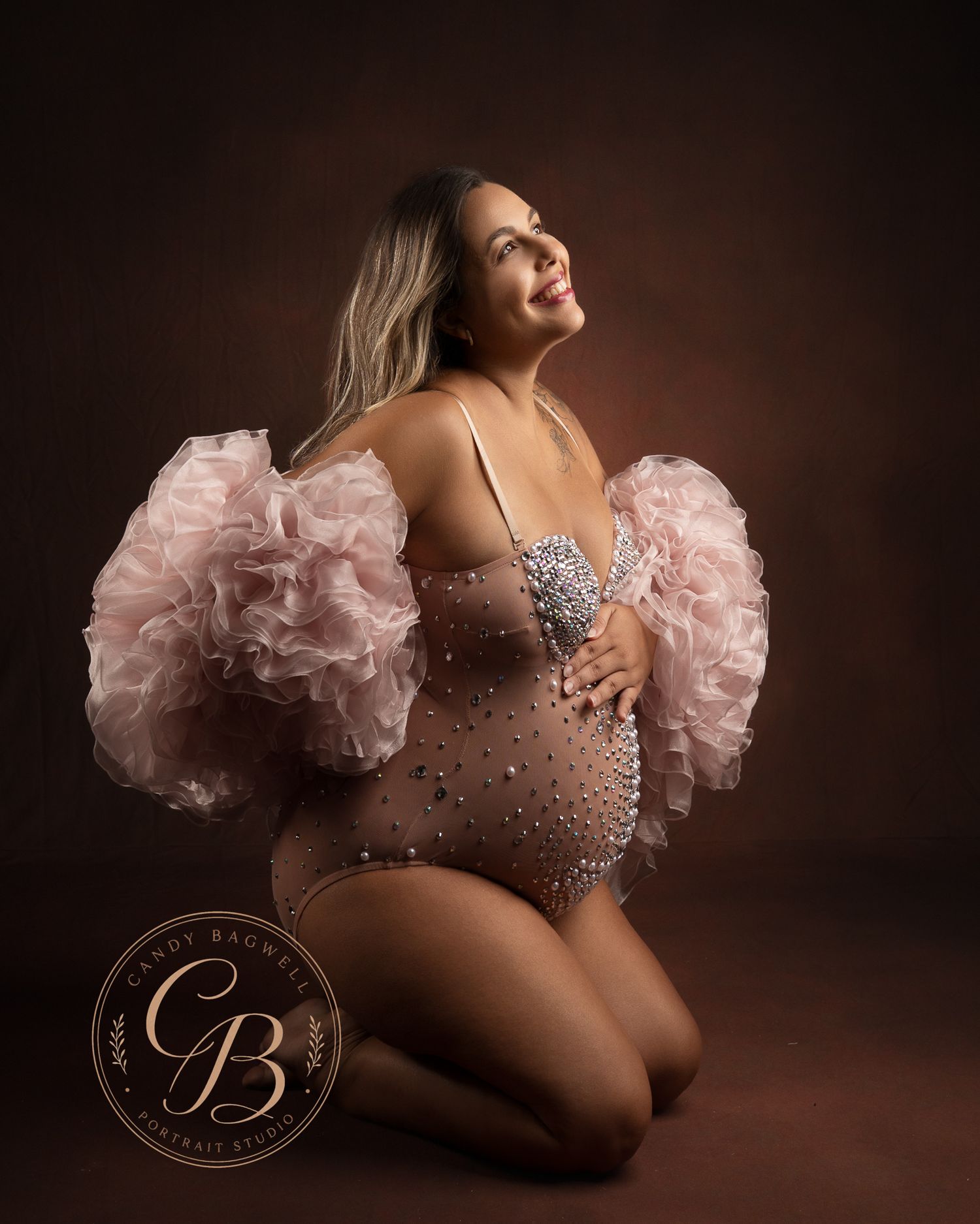 Pregnant woman in pink ruffled sheer bodysuit with pearl details, kneeling gracefully in a professional maternity photo session.