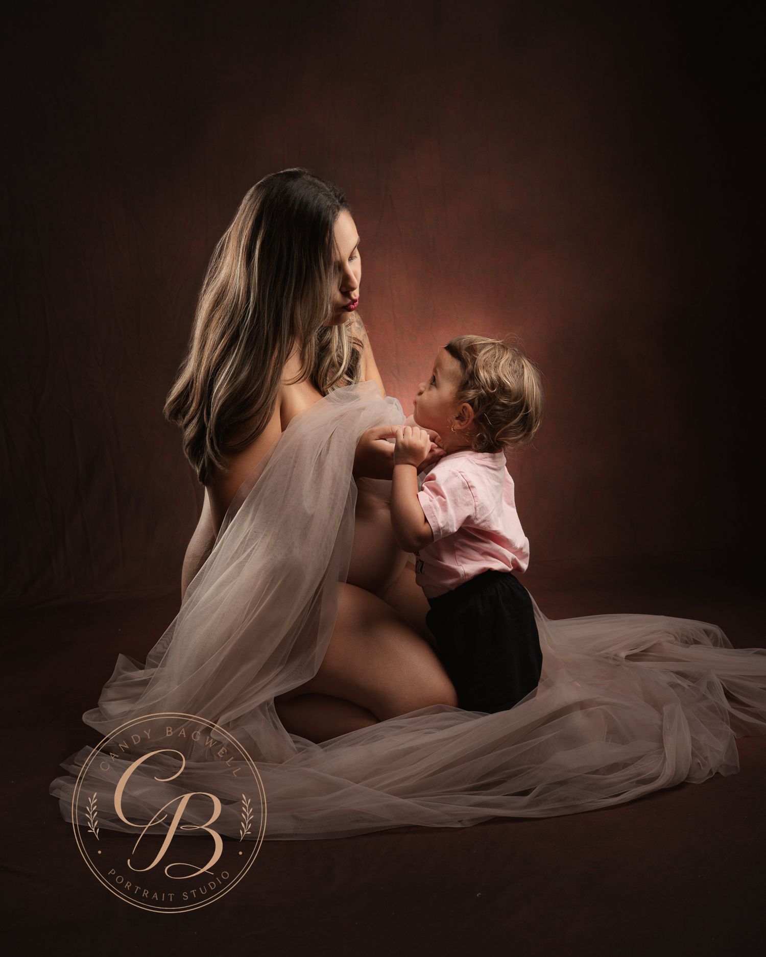 A pregnant woman in a flowing white tulle skirt sits on the floor with her young toddler in a warmly lit studio portrait.