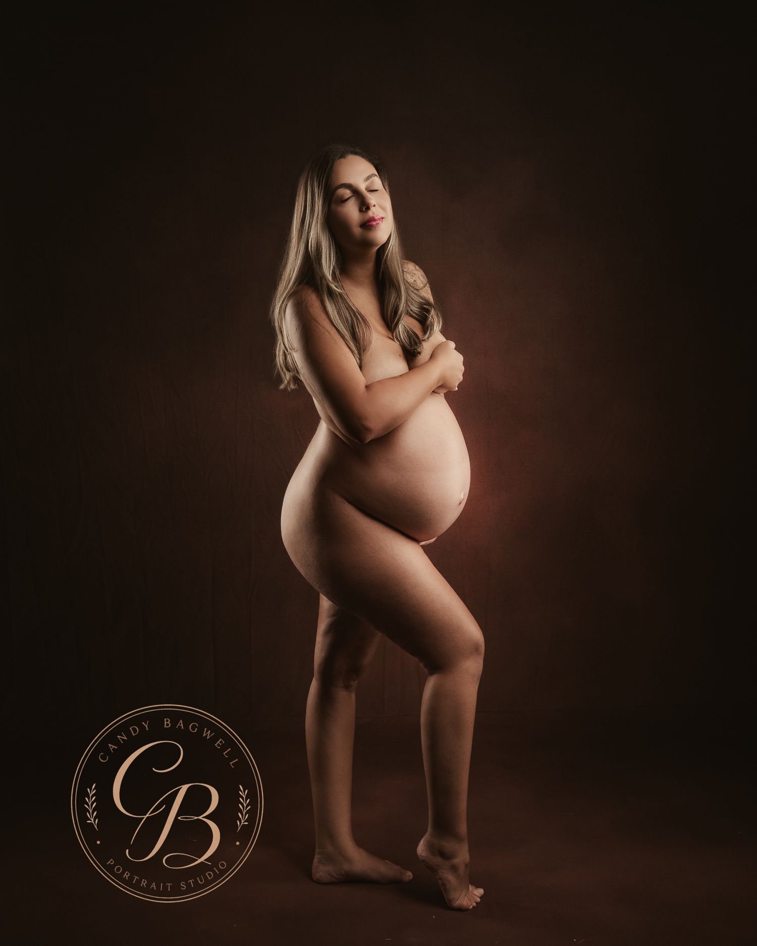 Elegant nude maternity portrait of a pregnant woman posing gracefully in a studio with warm brown background.