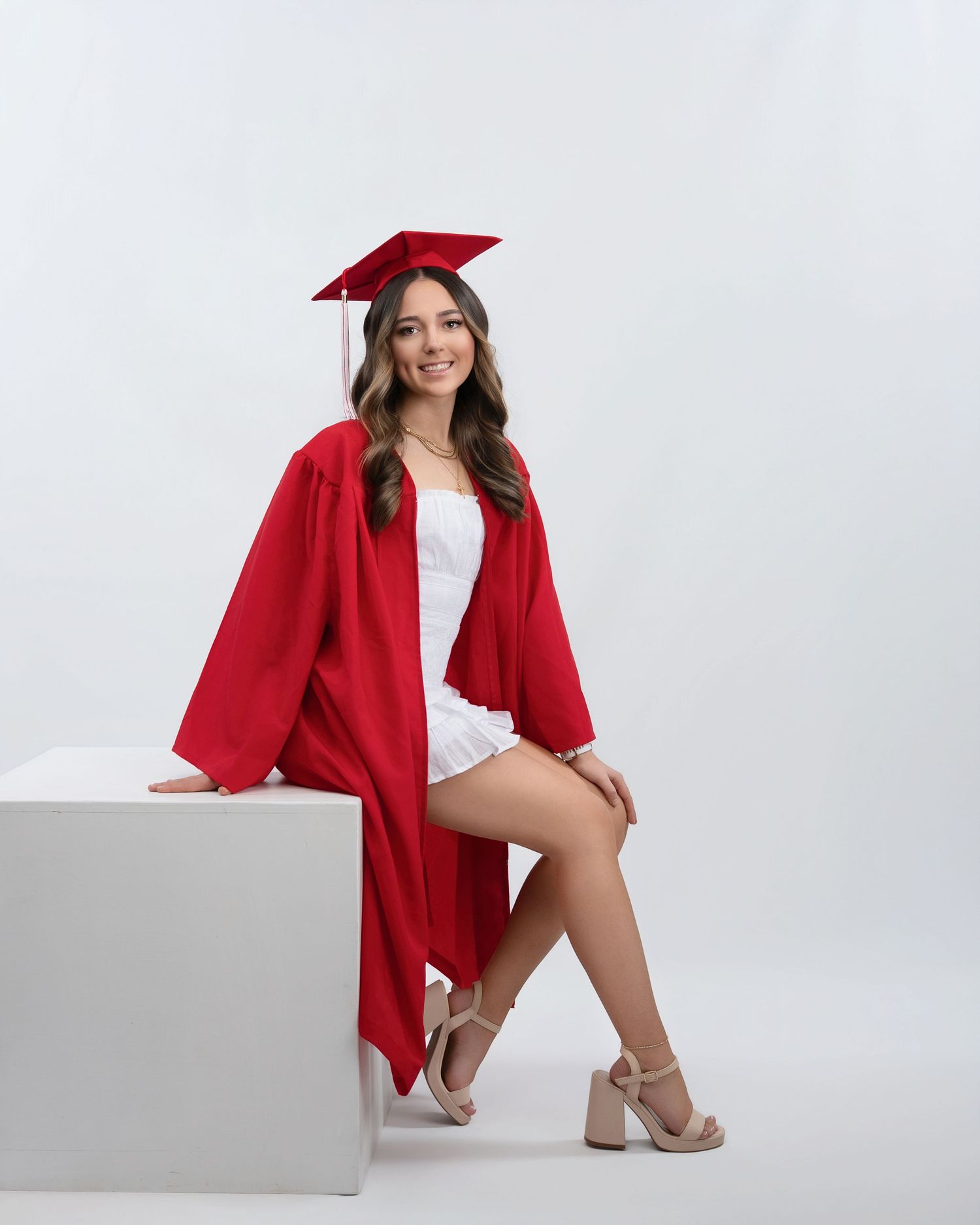 Smiling graduate in red cap and gown posing beside white cube in professional studio photo.