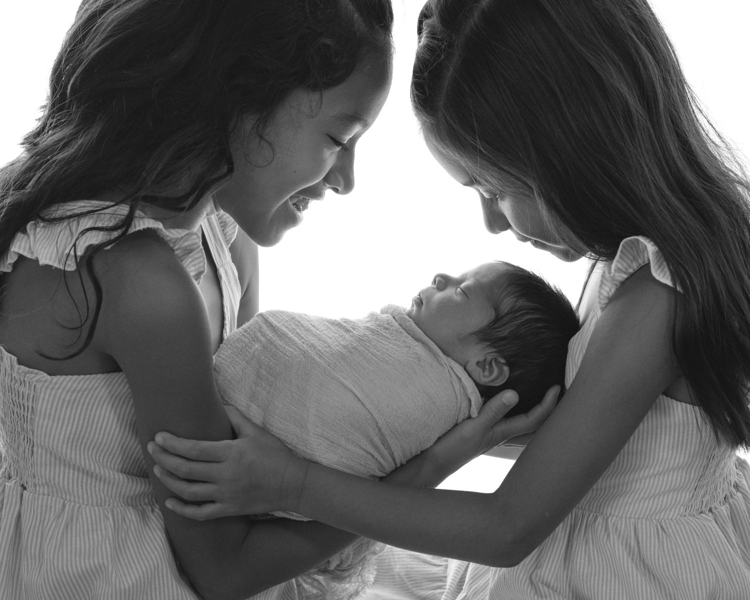 Black and white photo of two sisters smiling at swaddled newborn baby sibling they are holding.