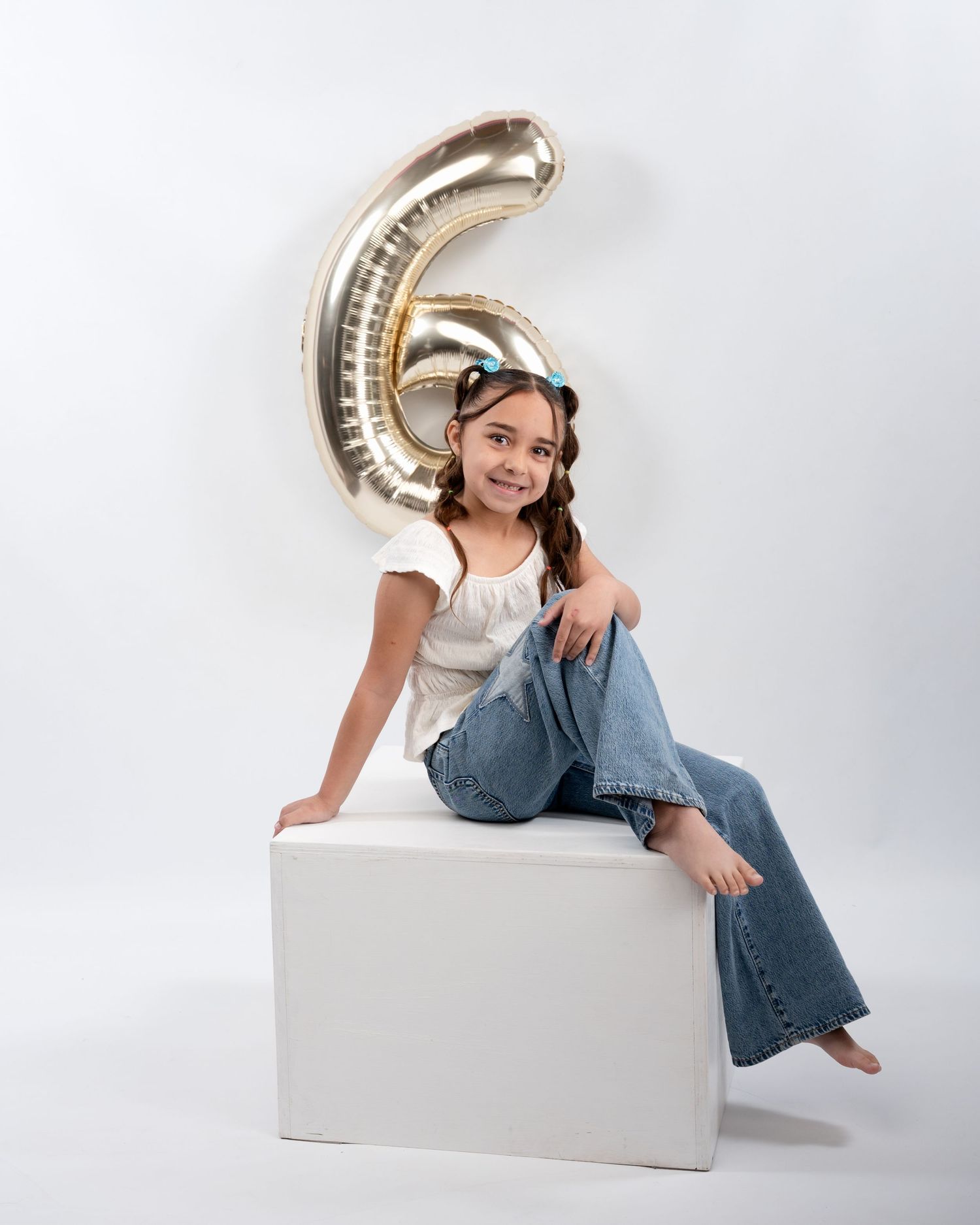 Smiling girl in white top and jeans sits on white cube beside large gold number 6 balloon.