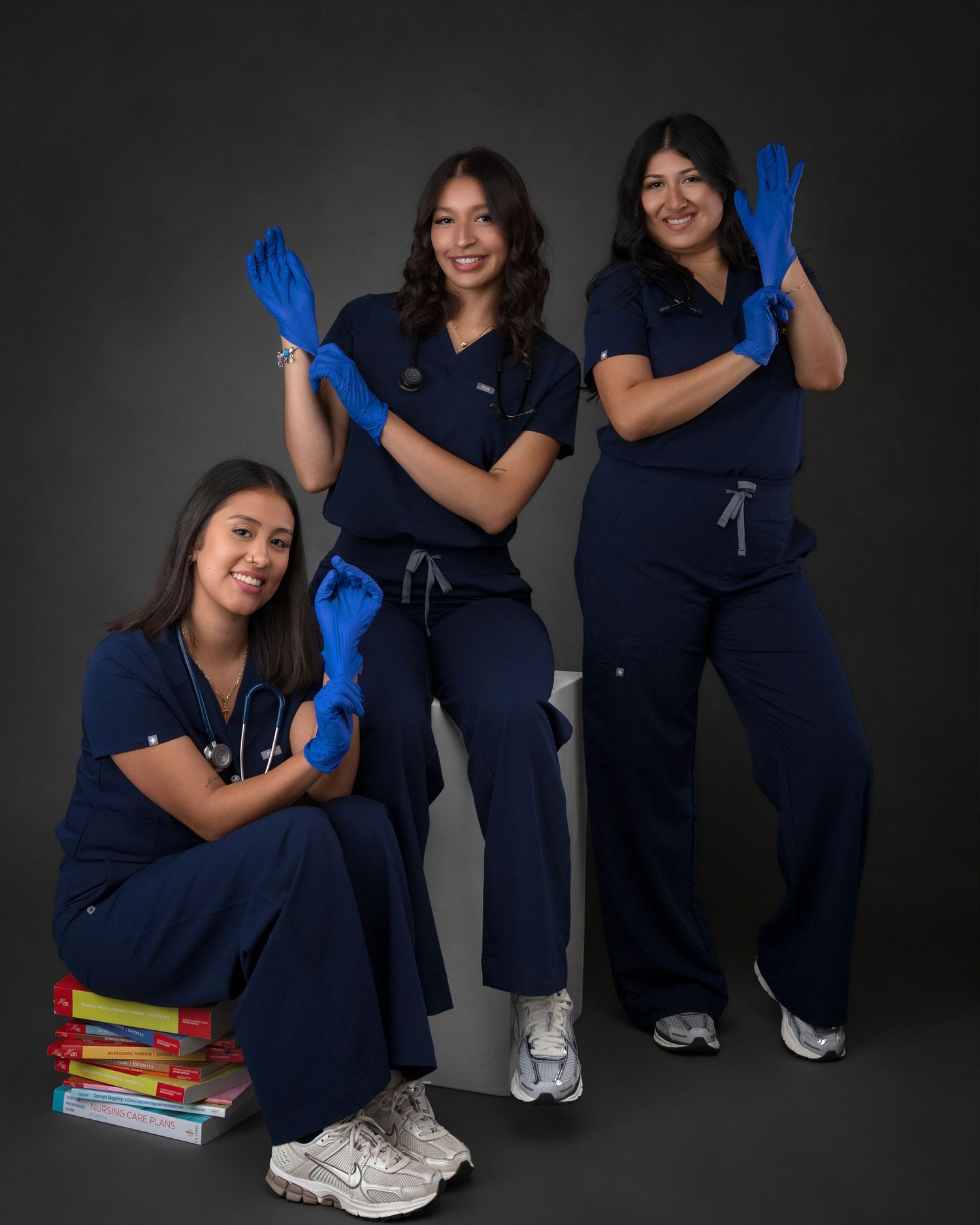 Three nursing graduates in navy scrubs pose playfully while putting on blue medical gloves.