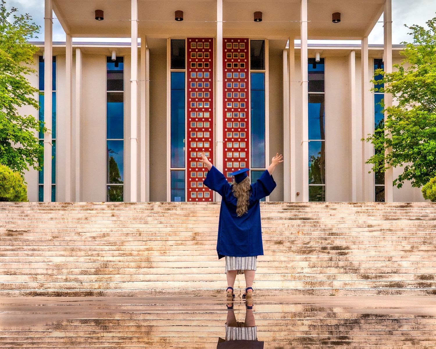 Graduate in blue cap and gown standing with arms raised before a grand building with ornate red doors.