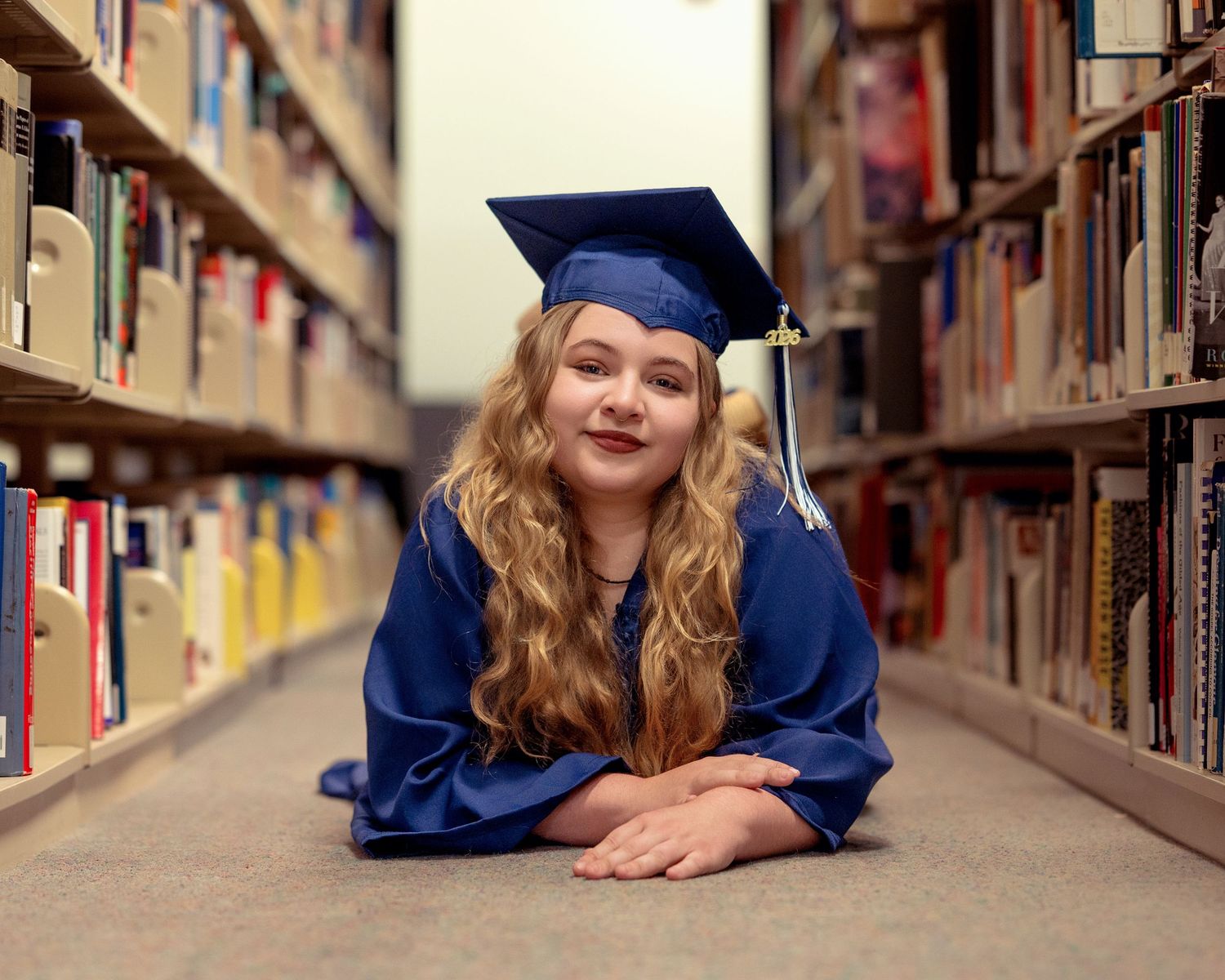 Young girl in blue graduation cap and gown lying on library floor between bookshelves, smiling at camera.