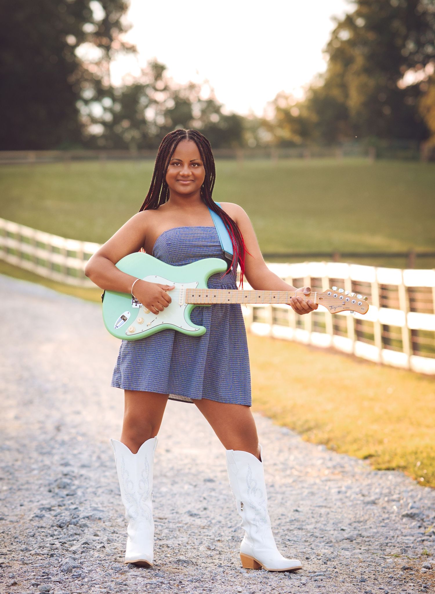 Young woman in denim dress and white cowboy boots holding a mint green electric guitar outdoors.