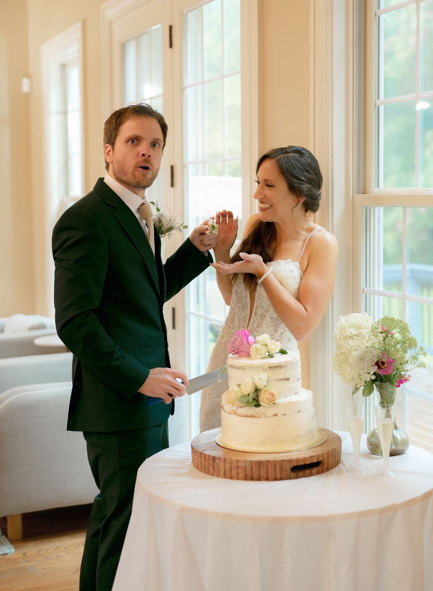 Groom playfully feeds bride wedding cake as they laugh together at their elegant indoor reception.