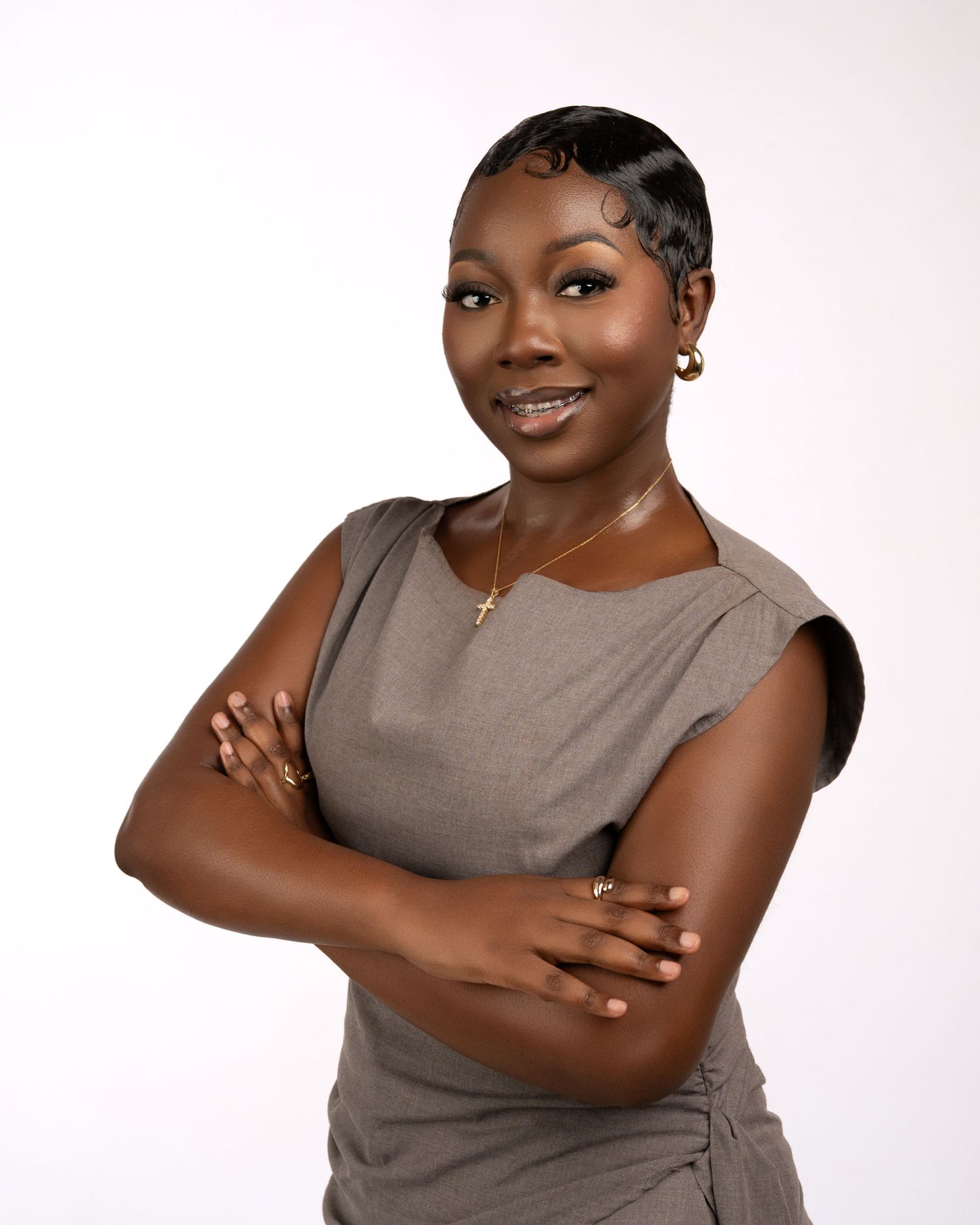 Professional woman in gray dress with arms crossed, wearing gold cross necklace, smiling confidently against white background.