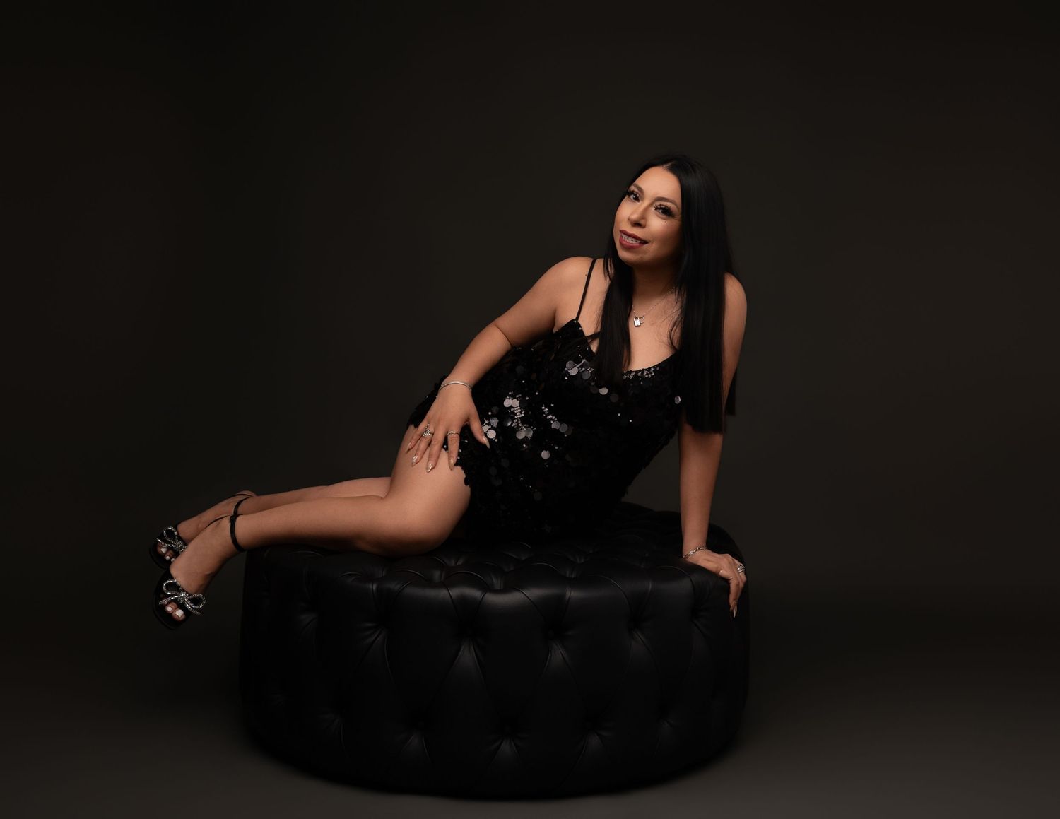Woman with long dark hair in a black sequined dress and strappy heels reclining on a tufted black leather ottoman in a dark studio portrait.