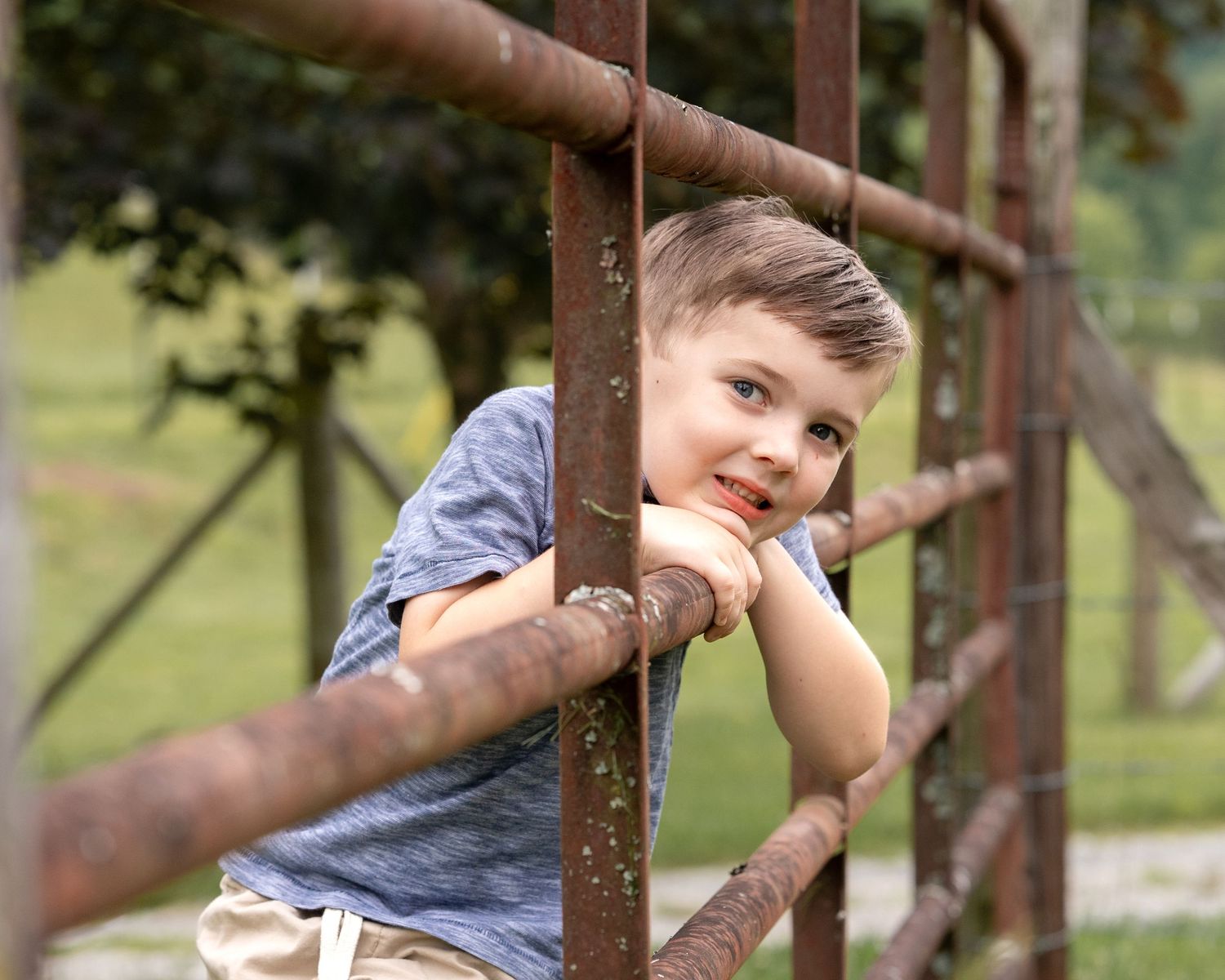 Smiling young boy leaning on rusty metal fence rails in a green outdoor setting.