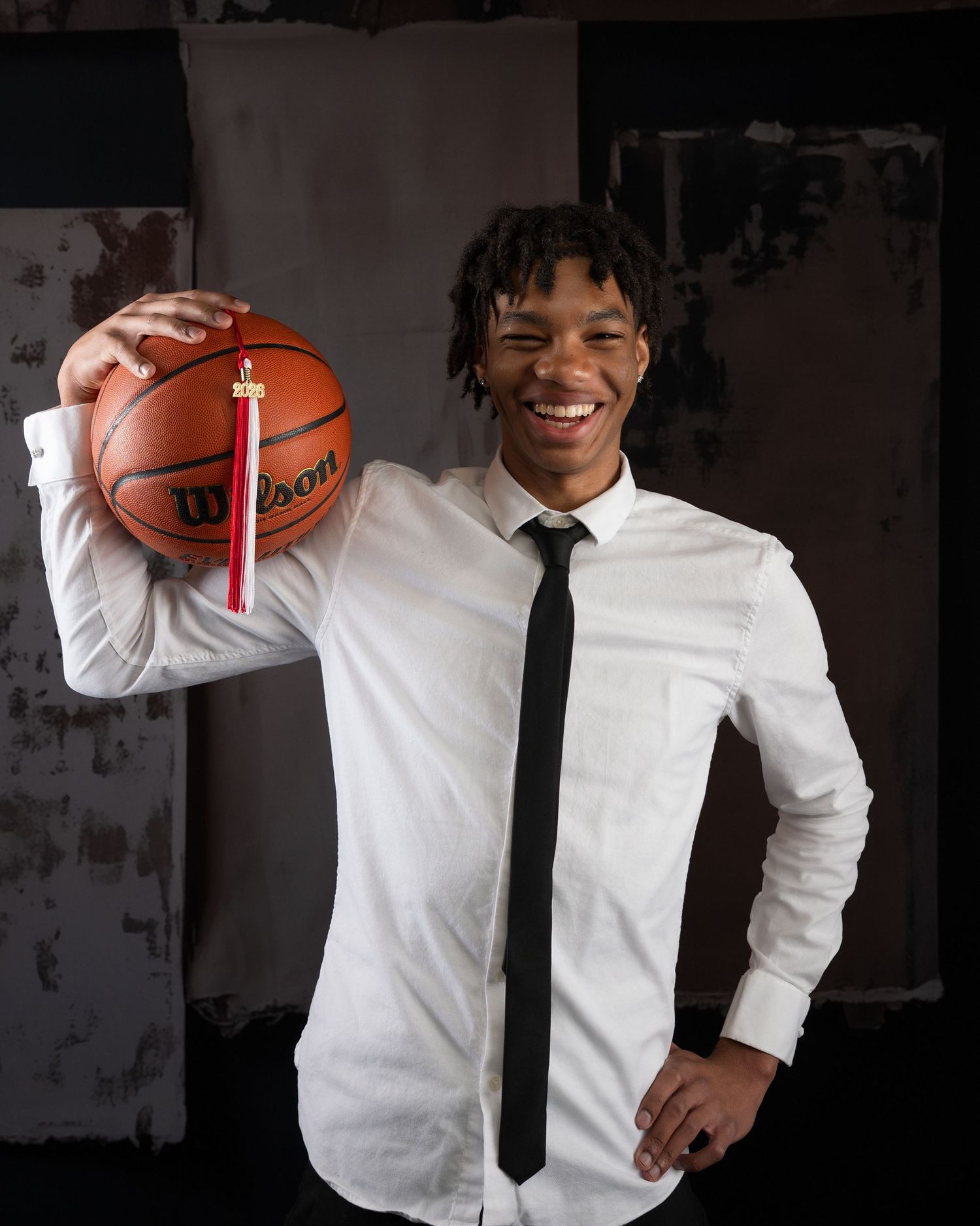 Smiling young man in white shirt and black tie holds Wilson basketball on shoulder in senior photo.