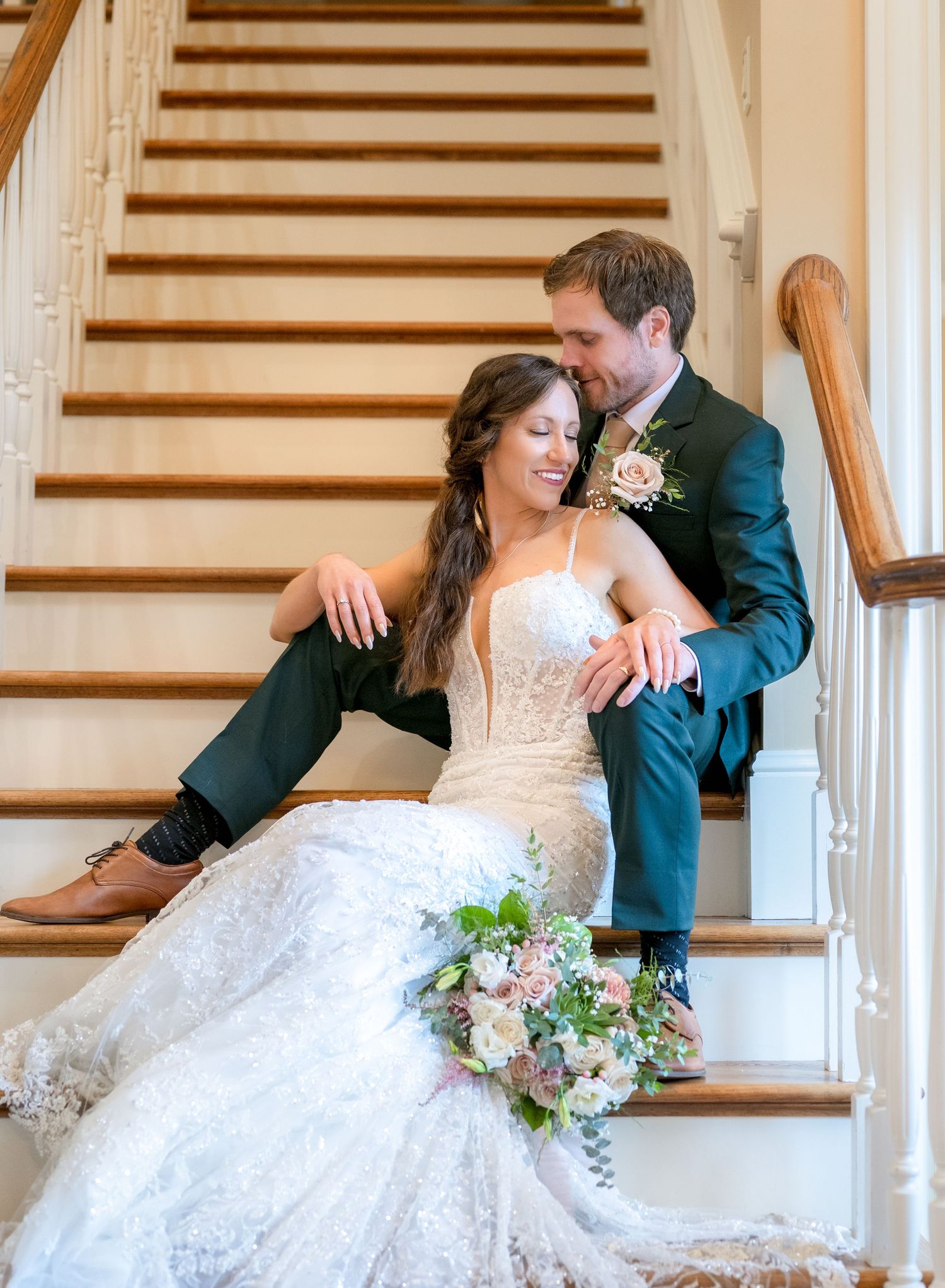 Bride in lace gown and groom in teal suit sit together on elegant staircase holding a pastel bouquet.
