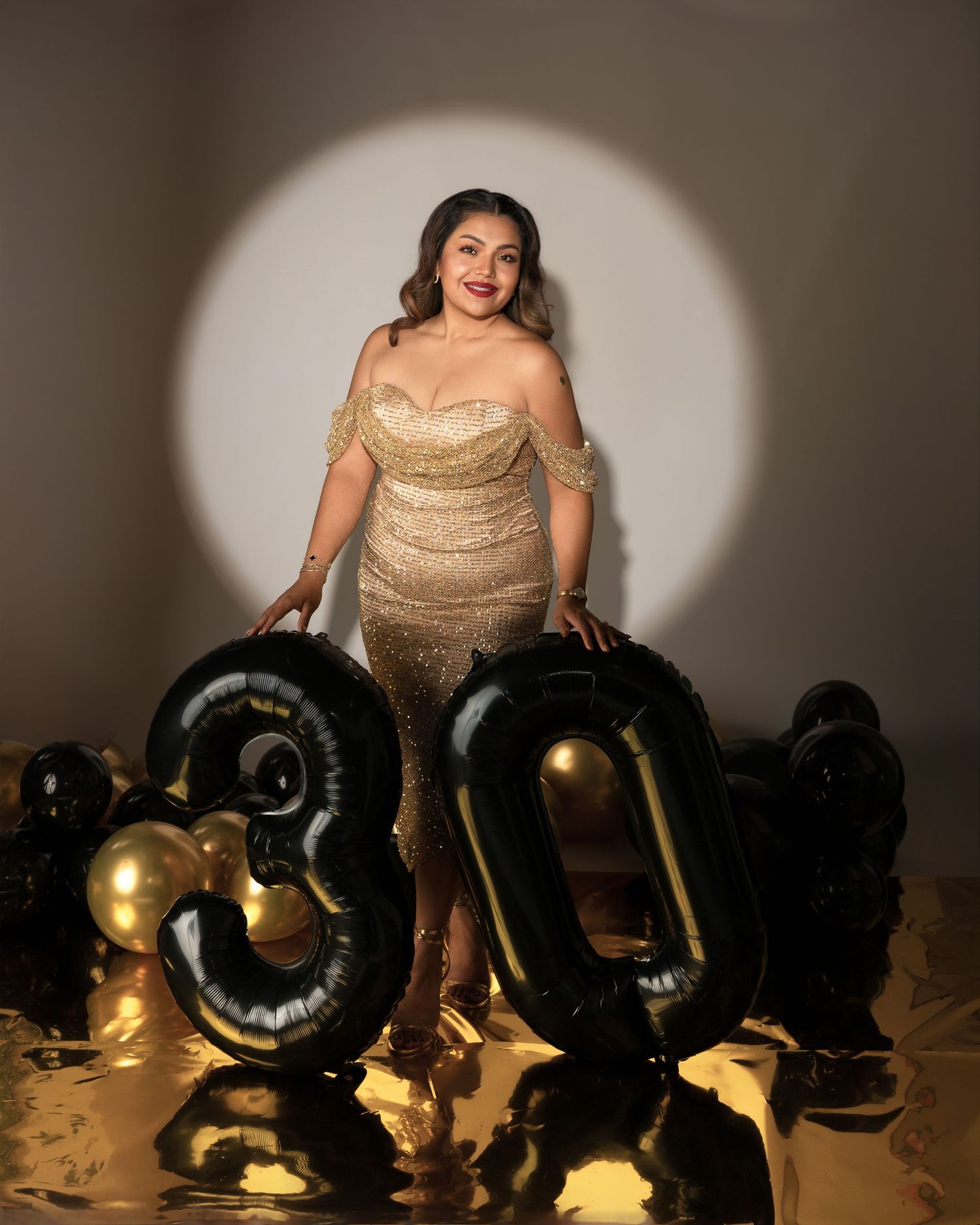 Woman in a sparkling gold off-shoulder gown poses under a spotlight beside black and gold balloons forming the number 30.