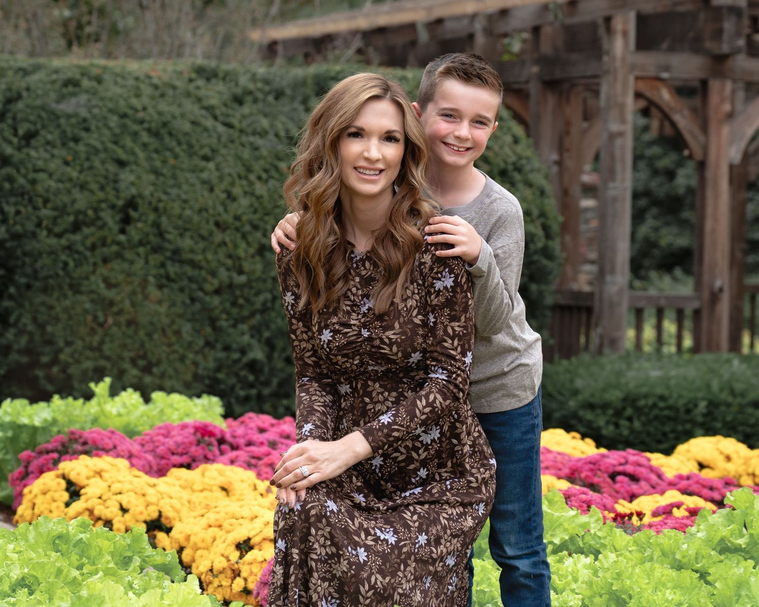 A brunette woman in a floral dress poses with a young boy in a vibrant garden.