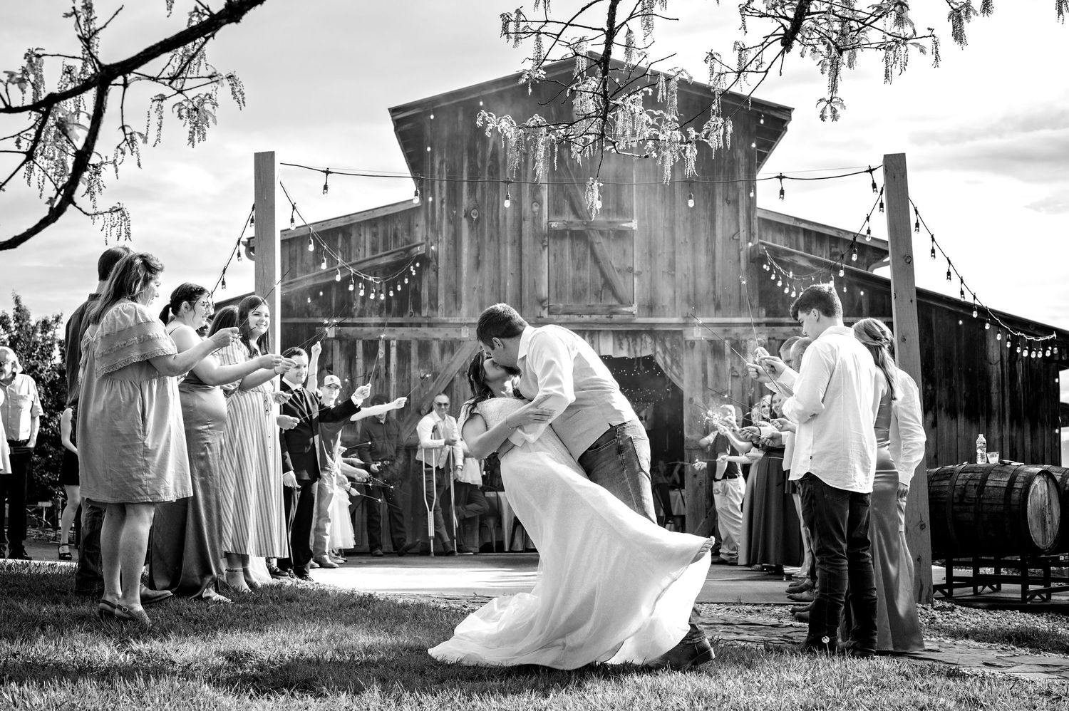 Black and white photo of groom dipping bride outside rustic barn wedding venue with guests.